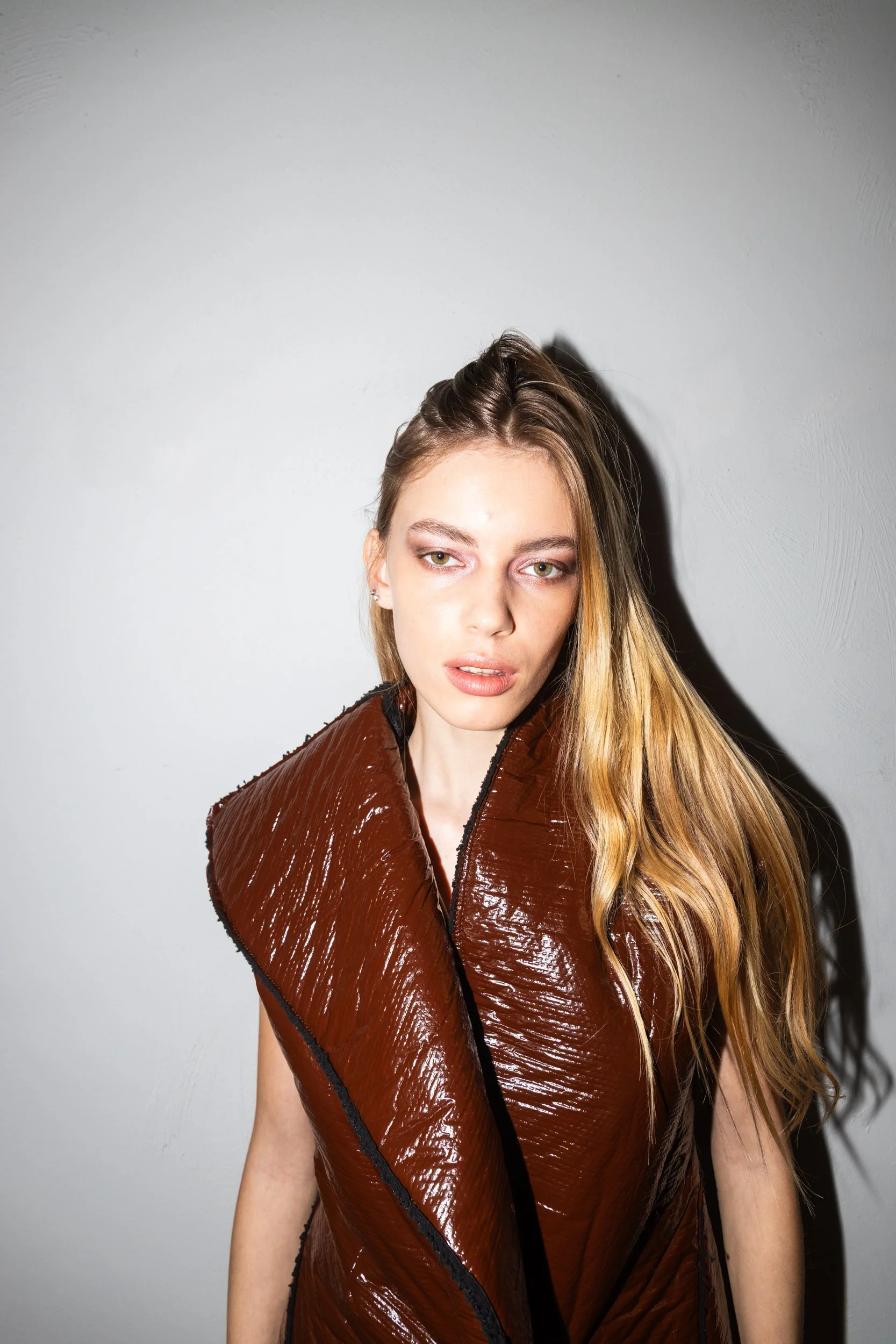 Young woman with long blonde hair, wearing a shiny brown vest, standing against a plain white wall, with a serious facial expression.