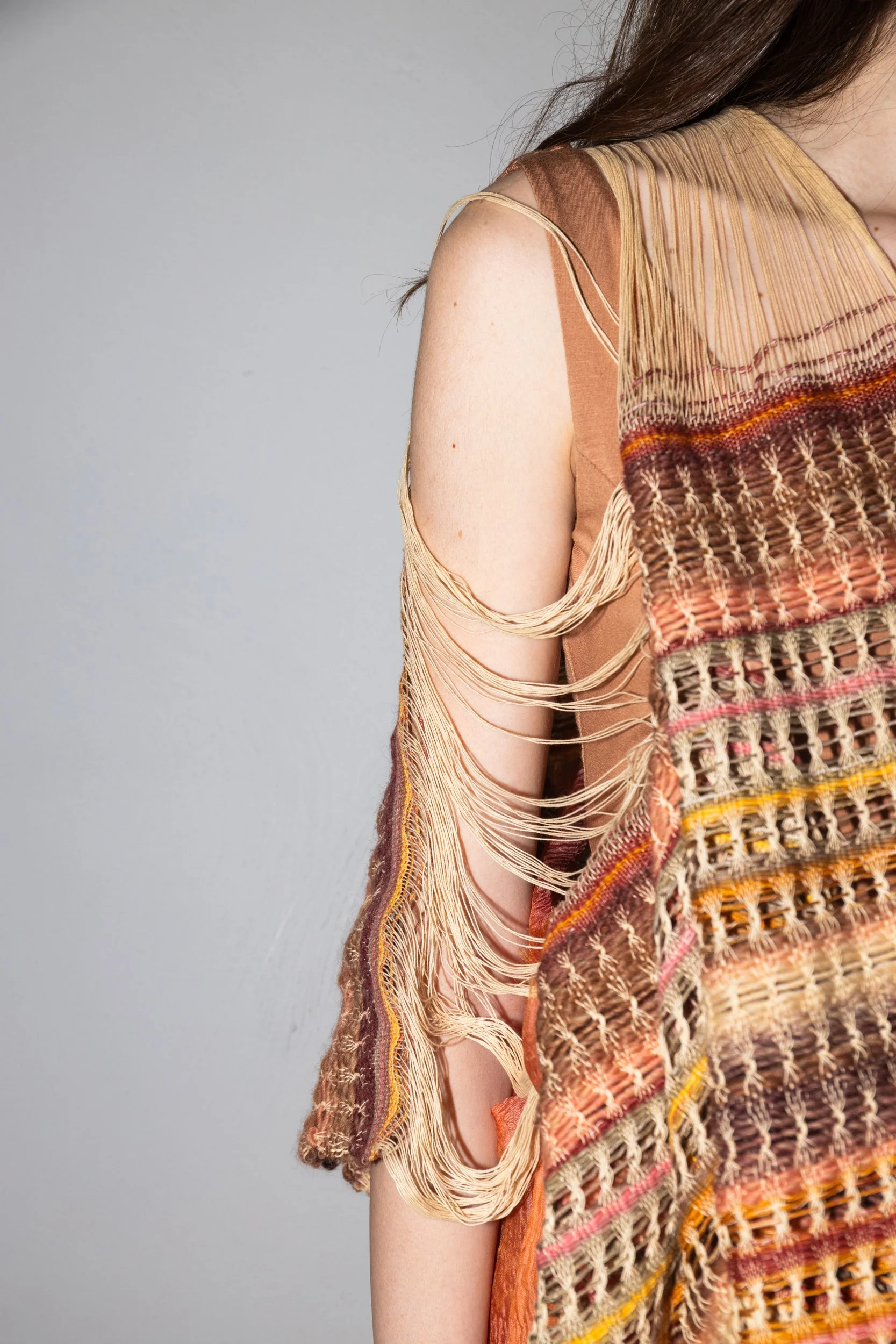 Close-up of a woman's shoulder and partial torso wearing a woven, fringed, multicolored top with strappy detailing on the arm against a plain background.