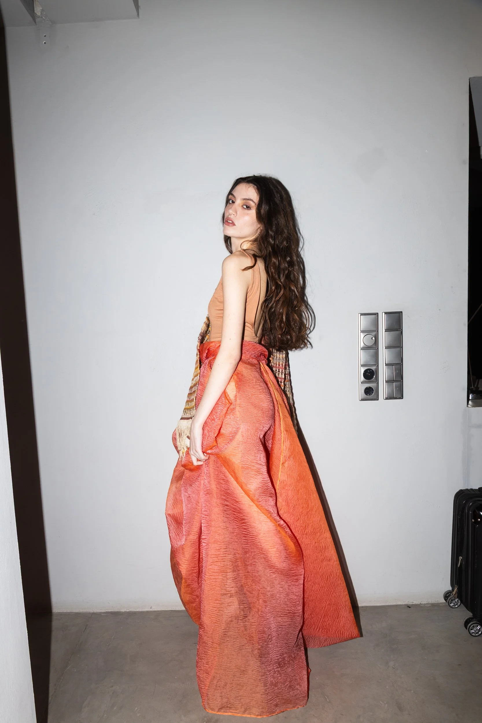 A woman with long, wavy brown hair posing against a plain gray wall, wearing a strapless peach-colored top and a large, flowing orange skirt.
