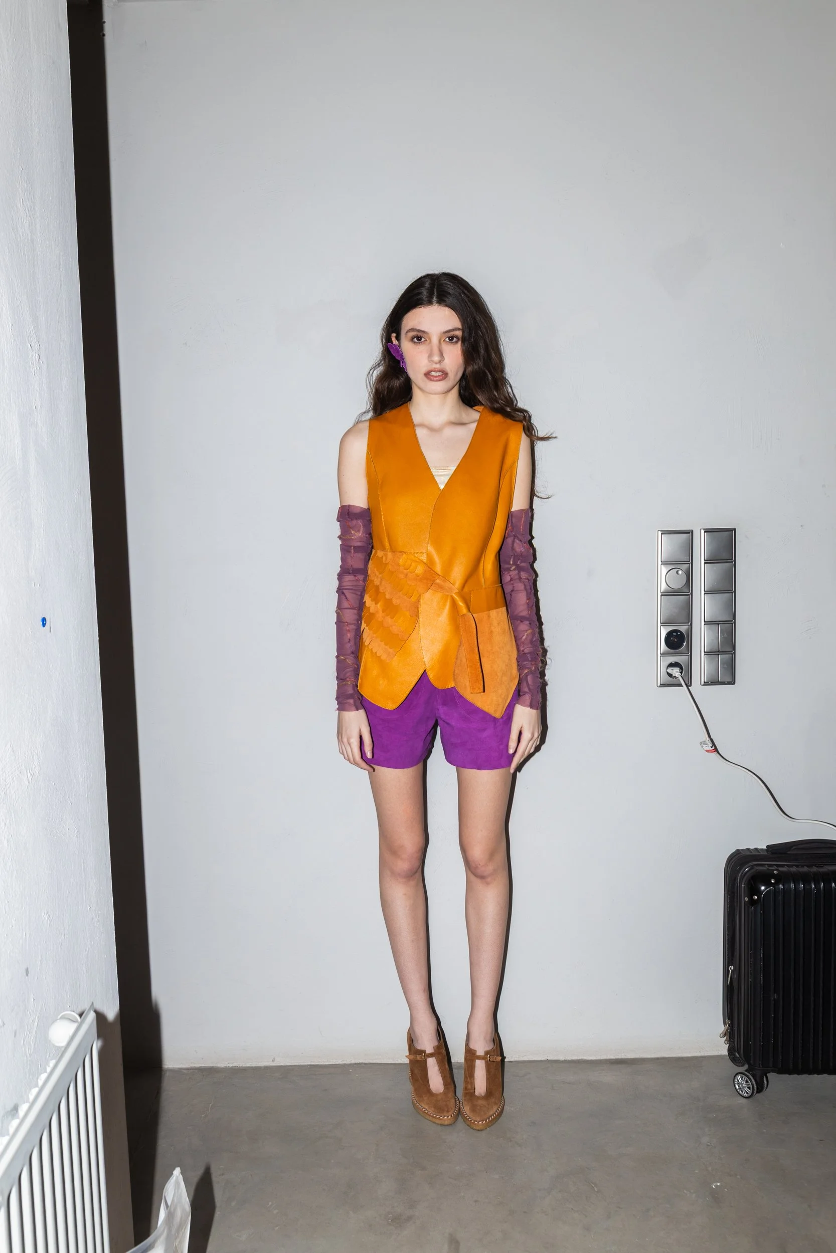 A woman standing against a plain wall, wearing a mustard orange vest, purple shorts, sheer purple sleeves, and brown high-heeled shoes.