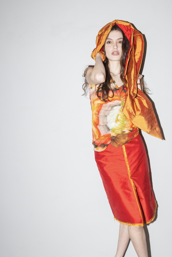 A woman with long dark hair wearing a colorful top, a red skirt, and a large orange and red hat standing against a plain white wall.