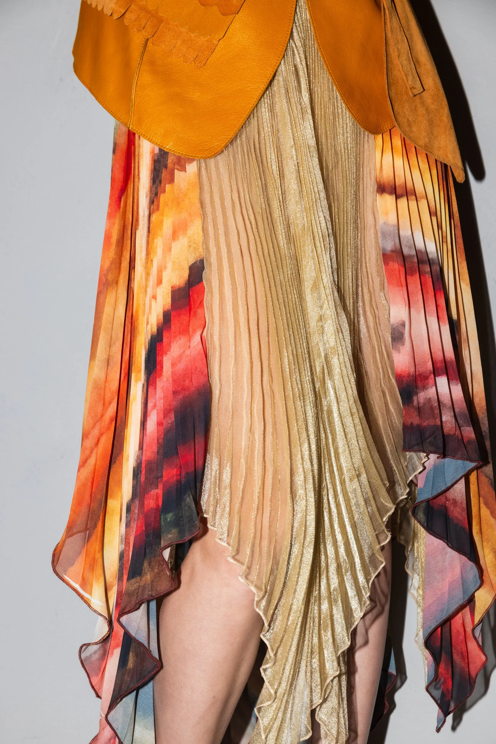 Close-up of a person wearing a multicolored, pleated, metallic dress and an orange leather jacket.