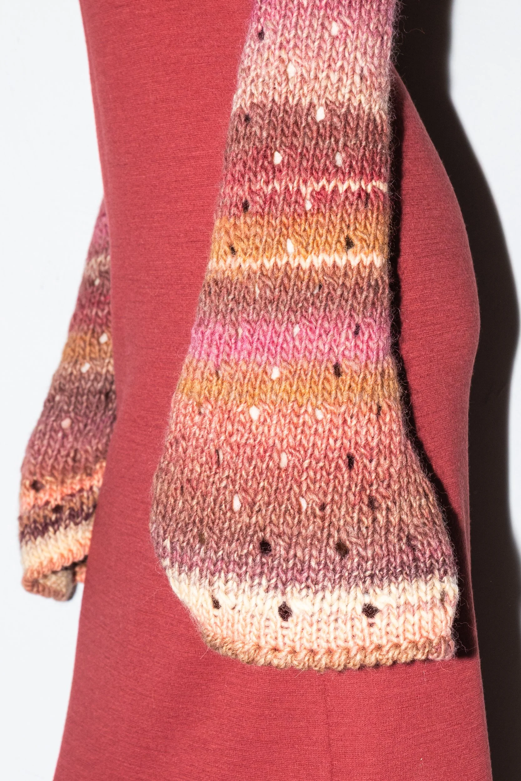 Close-up of a multicolored, hand-knit sleeve with shades of pink, orange, and brown, worn over a maroon long-sleeve shirt.
