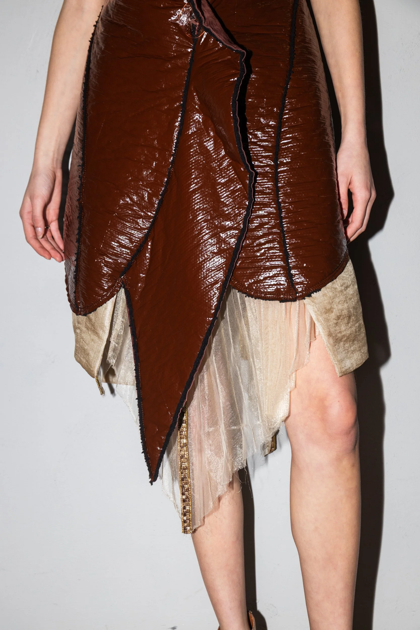 A person wearing a layered outfit with a shiny brown asymmetrical leather top over a beige skirt with sheer fabric details.