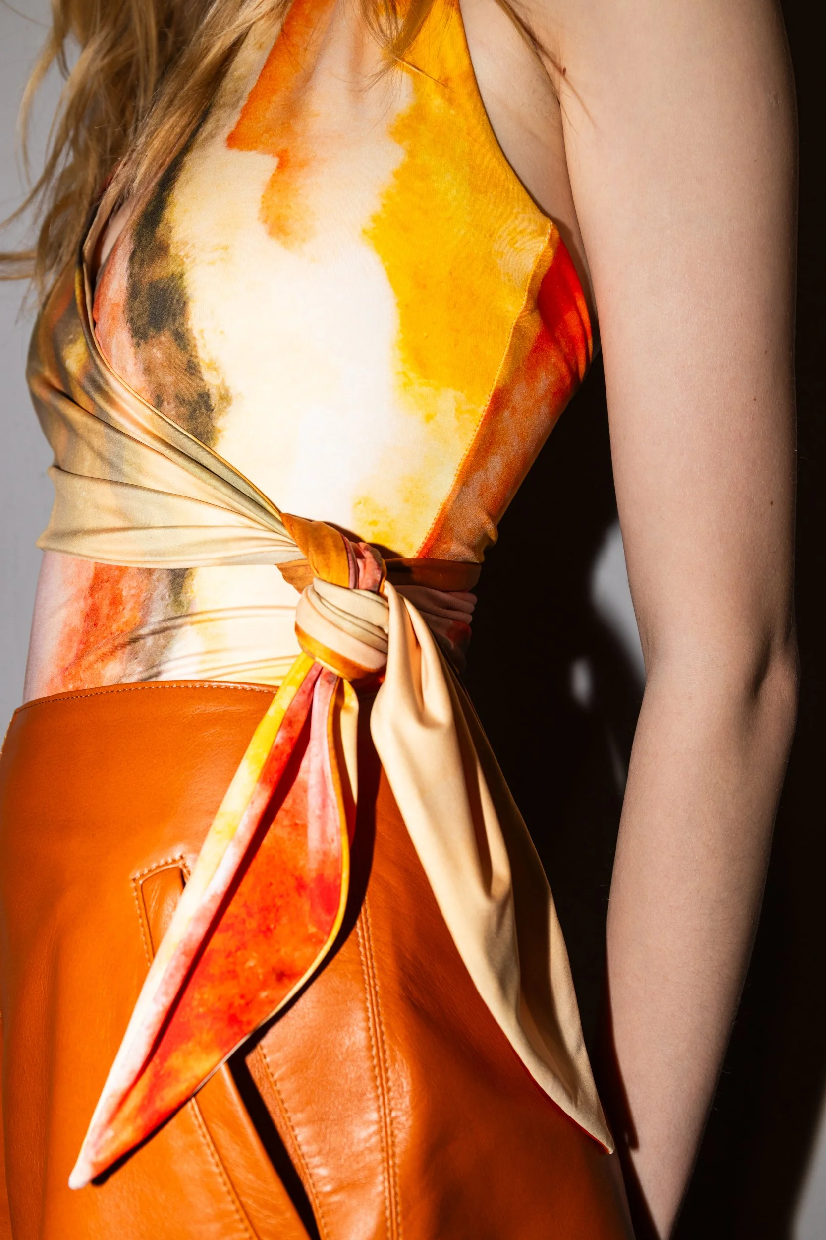 Close-up of a woman wearing an orange and beige patterned sleeveless top with a tie at the waist, and orange leather pants.