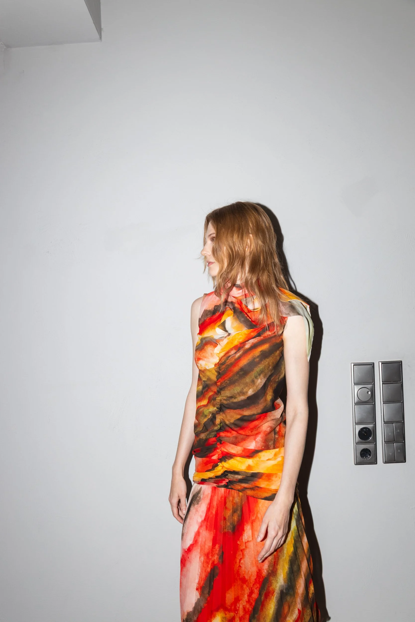 A woman with shoulder-length red hair wearing a sleeveless dress with a fiery, abstract pattern in red, orange, yellow, and brown, standing against a plain white wall.