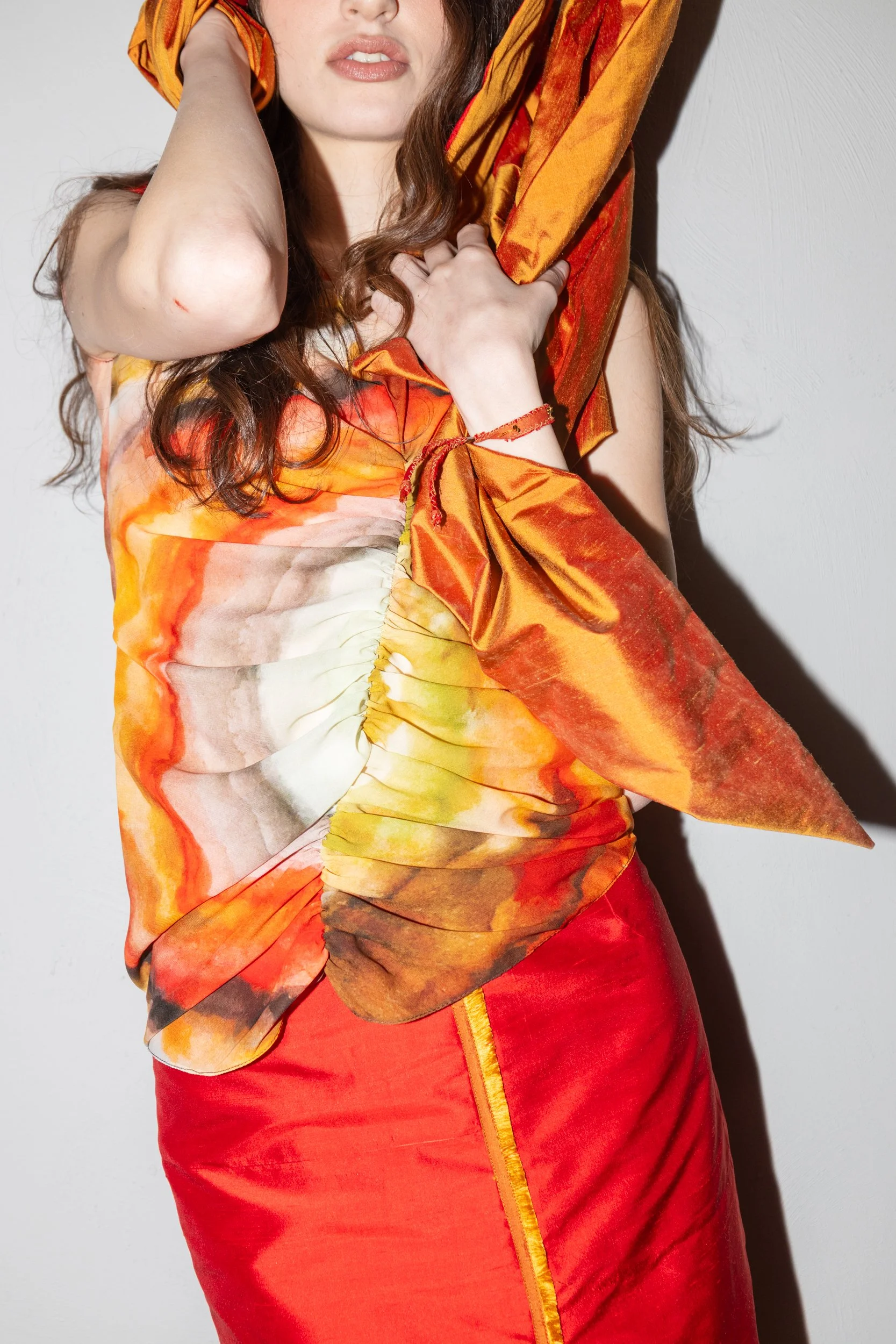 A woman wearing a colorful, abstract patterned top in shades of orange, yellow, brown, and cream, paired with a bright red skirt with orange piping, is posing against a plain white wall. She has long, wavy brown hair and is holding a yellow-orange sc