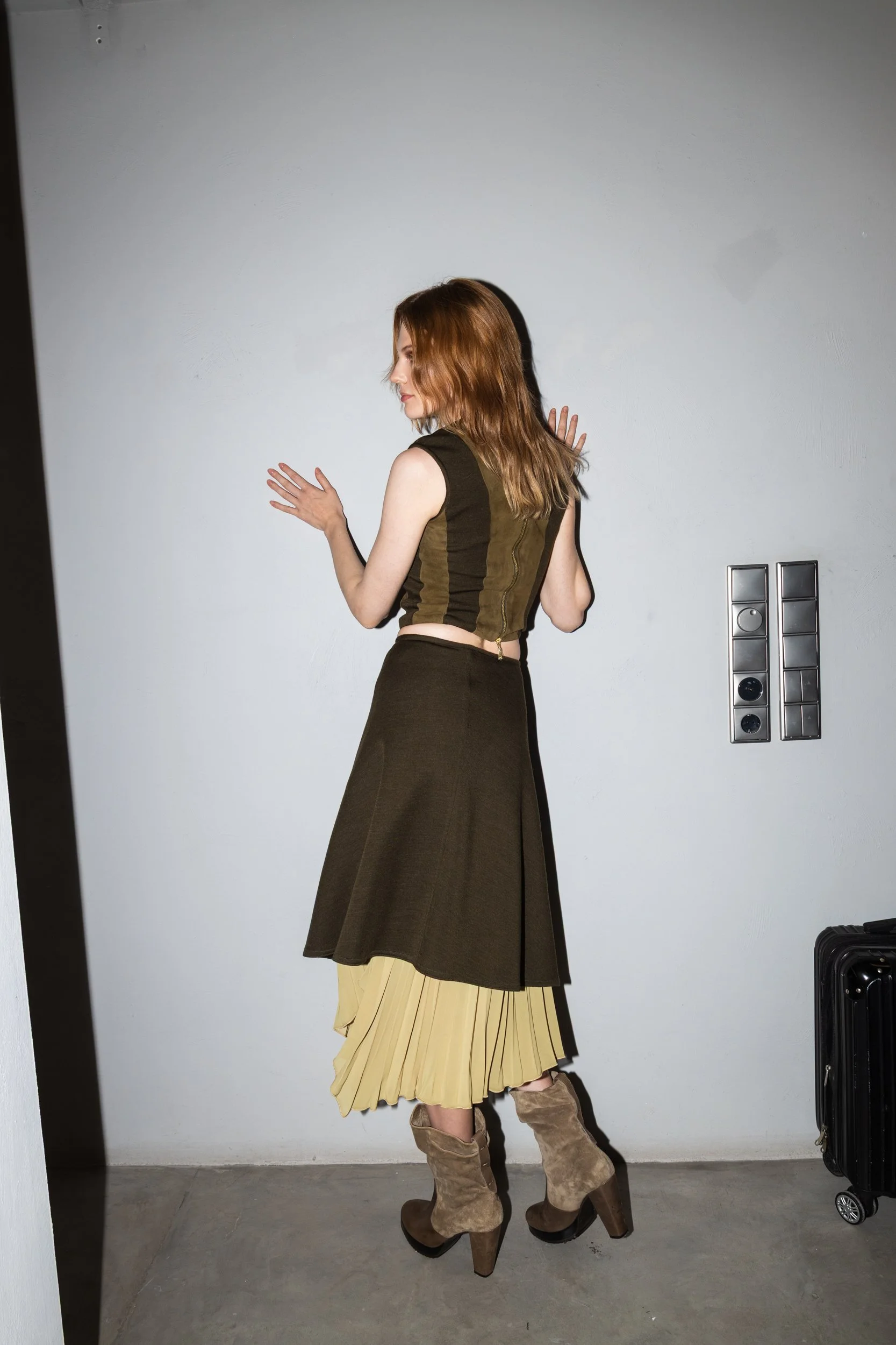 A woman standing against a plain white wall with a luggage cart nearby, wearing a sleeveless top, layered skirt, and high-heeled boots.