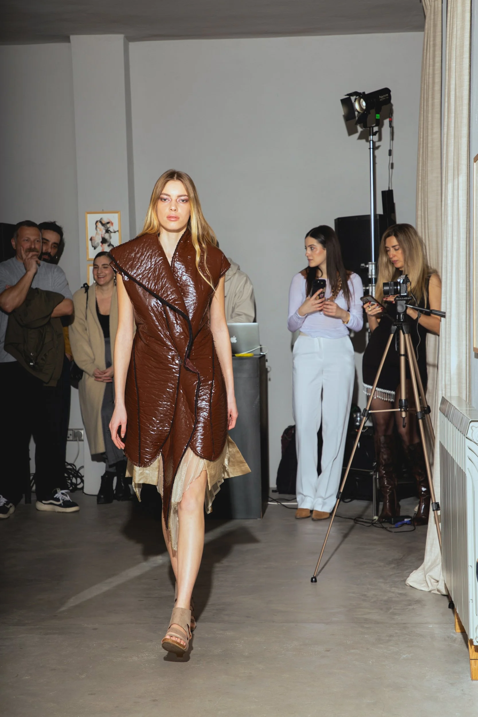 A fashion model walking down the runway during a fashion show, wearing a brown textured sleeveless coat with a layered beige skirt and beige sandals, with backstage crew and photographers in the background.