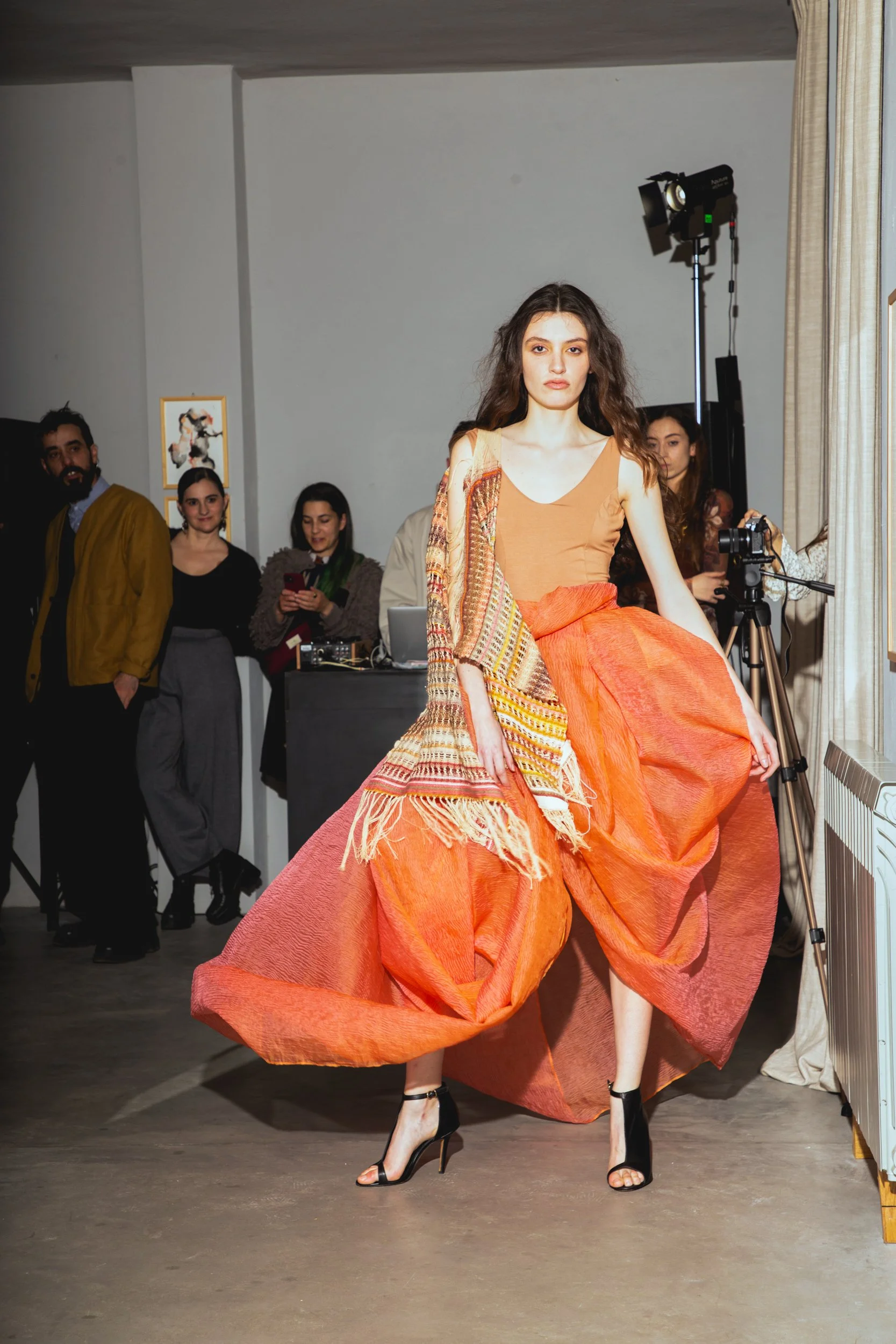 Fashion model walking in a runway show, wearing an orange and beige outfit with high heels, inside a room with onlookers in the background.
