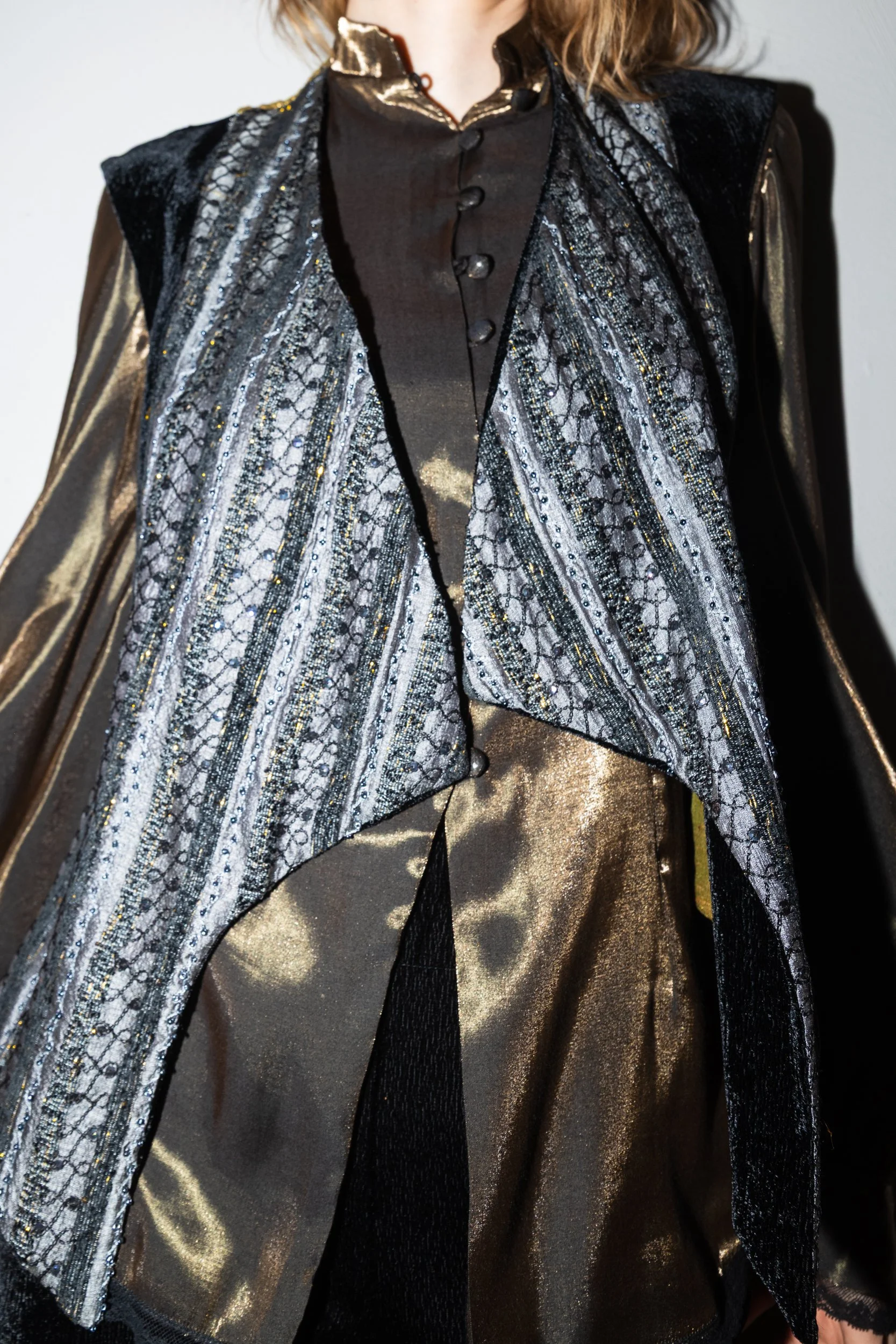 Close-up of a mannequin wearing a black satin blouse with buttons, layered under a metallic gold and patterned black-and-silver cape with textured detailing.