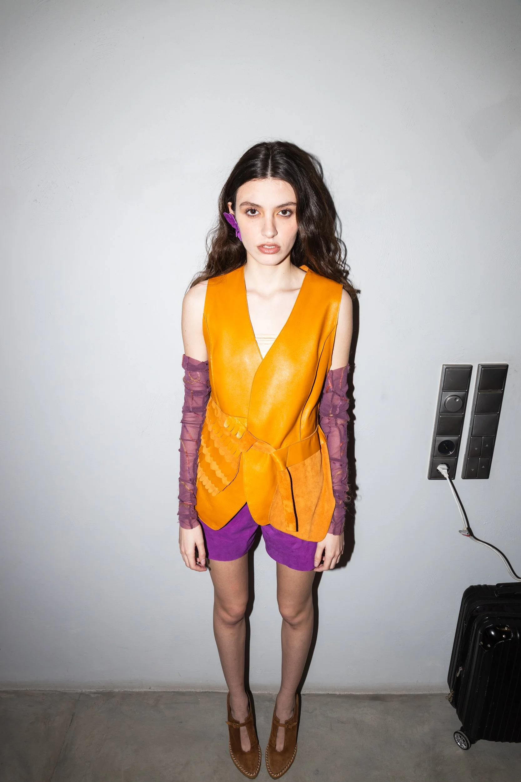 A young woman with long, curly brown hair stands against a plain white wall. She wears an orange sleeveless top with layered fabric and purple shorts. She has a purple hair clip and is wearing brown heeled shoes. A black rolling suitcase is seen on t