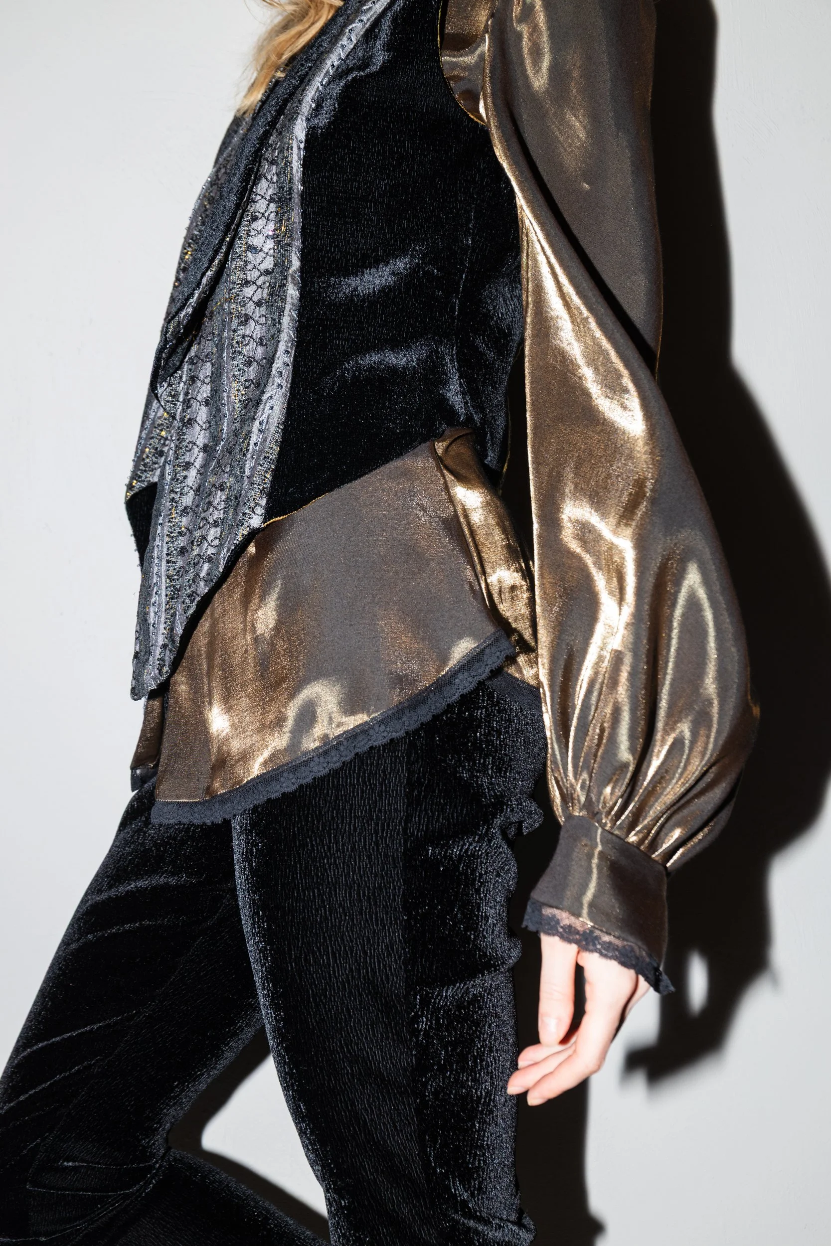 Close-up of a woman wearing a black velvet top and metallic gold and sheer jacket with puffed sleeves, standing against a white background.