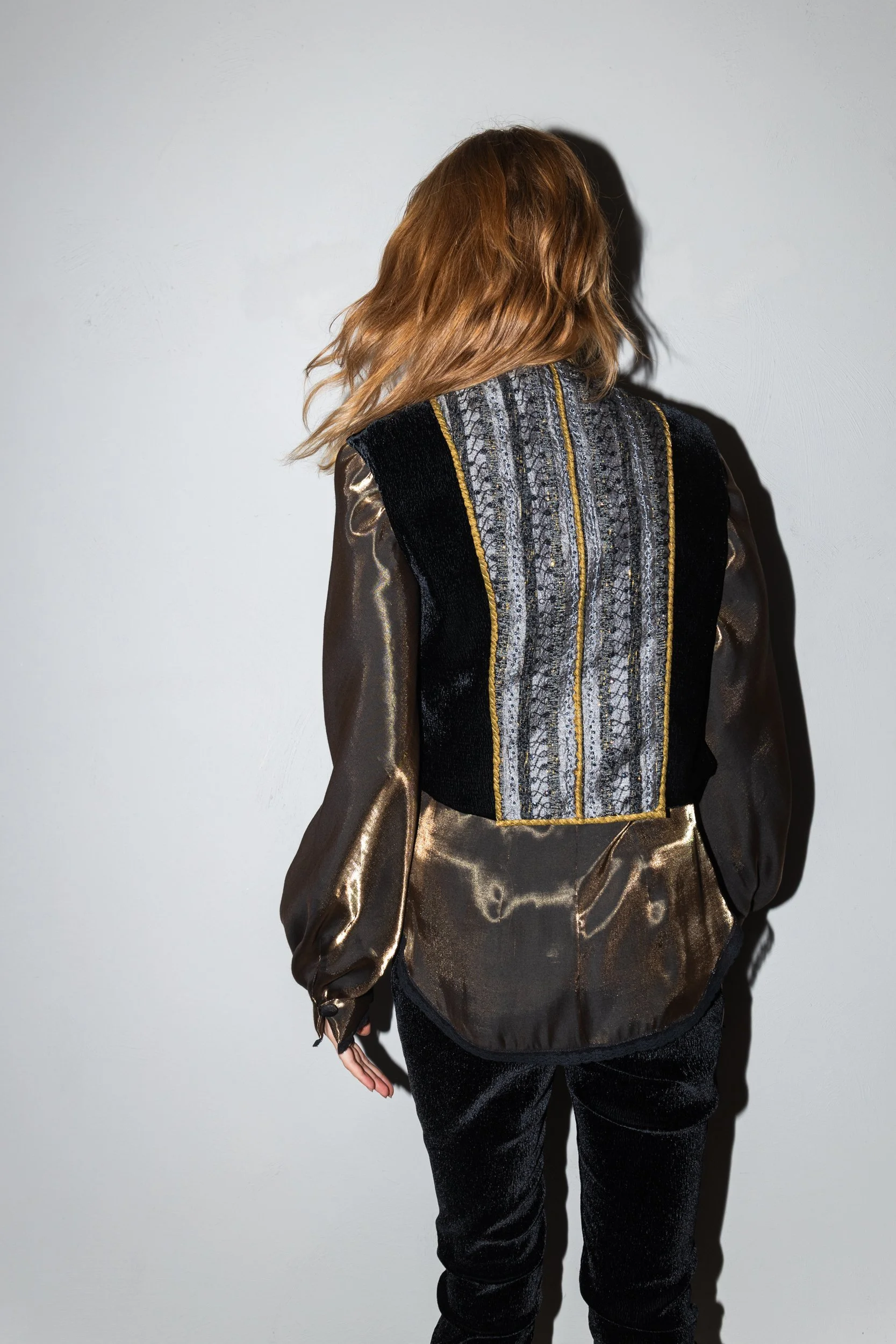 Woman with shoulder-length brown hair wearing a metallic shirt and a vest with intricate embroidery, facing away from the camera against a plain white wall.