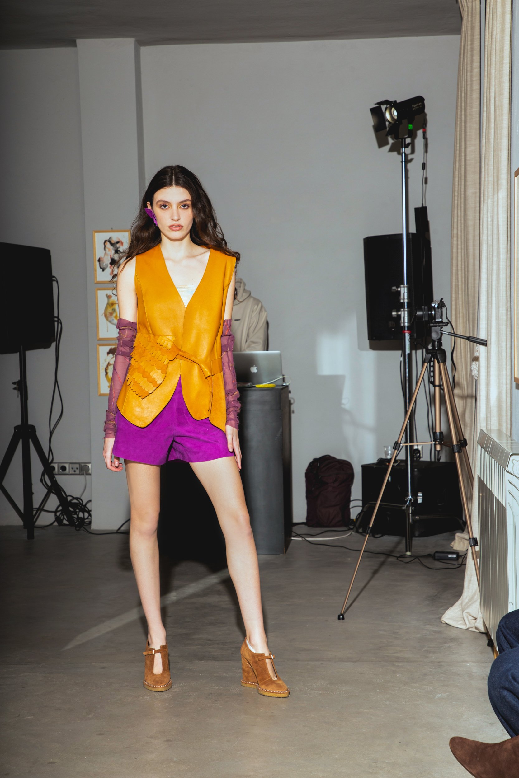 A young woman posing in fashionable clothing in a photography studio, wearing a mustard yellow vest, purple shorts, and brown high-heeled shoes with heels, with professional lighting and equipment in the background.