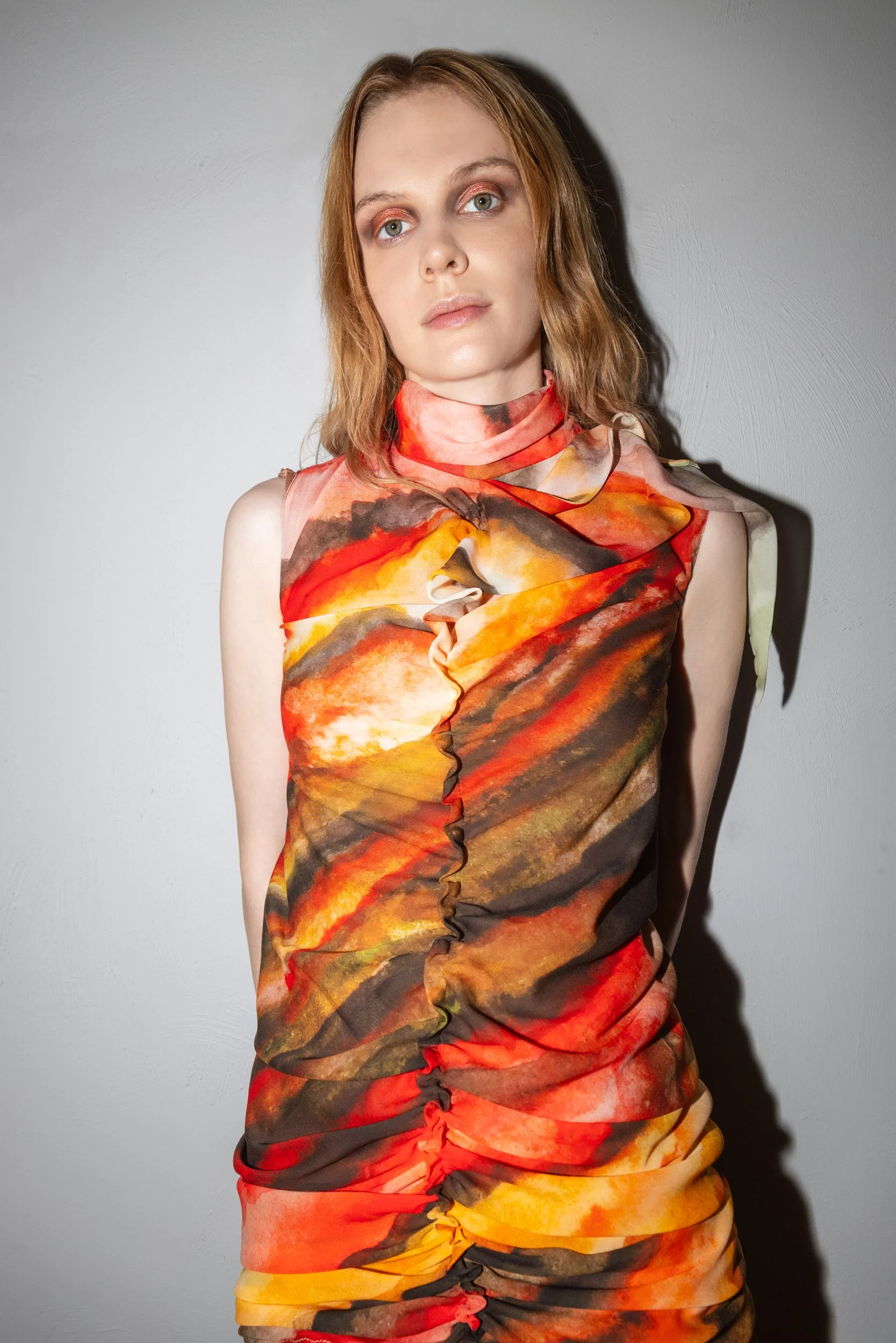 Woman with shoulder-length hair wearing a sleeveless dress with a fiery sunset pattern, standing against a gray wall.