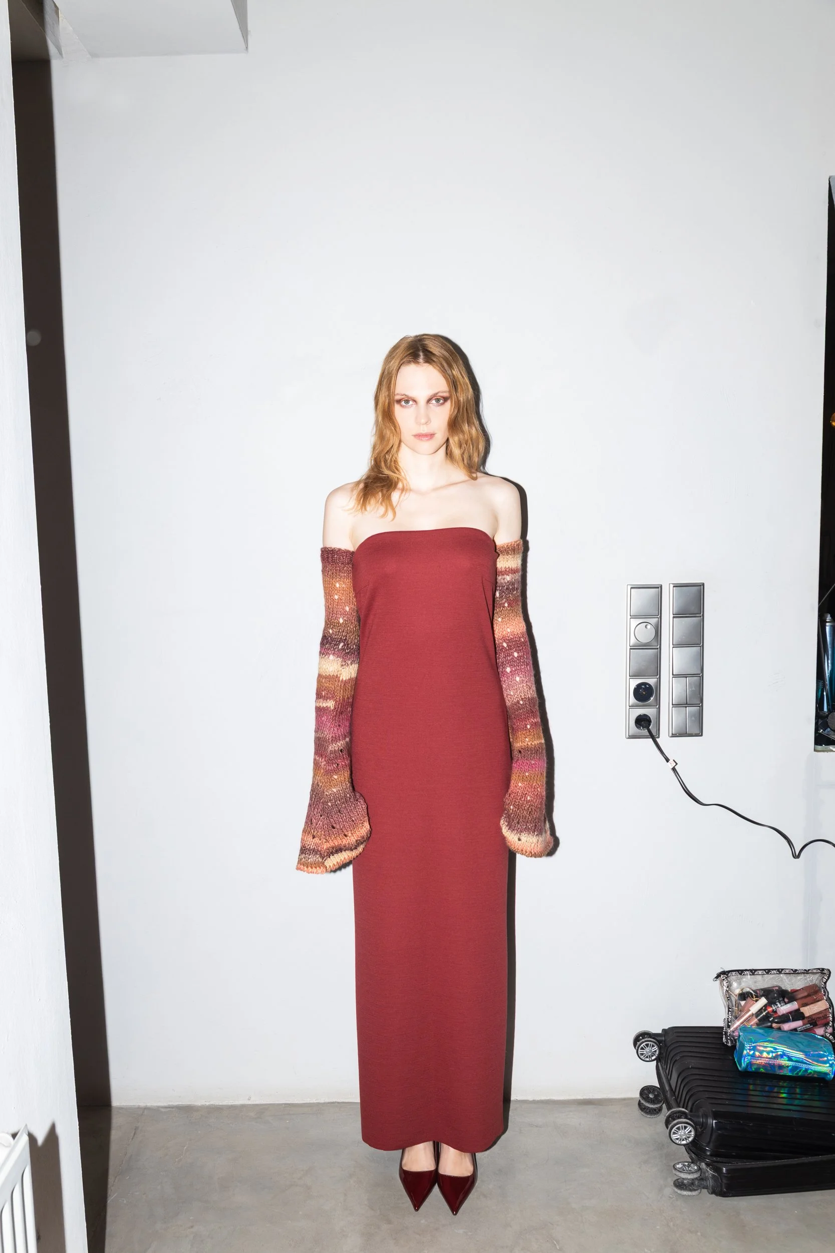 A woman with red hair standing against a white wall, wearing a strapless maroon dress, multicolored knit sleeves, and red high heels.