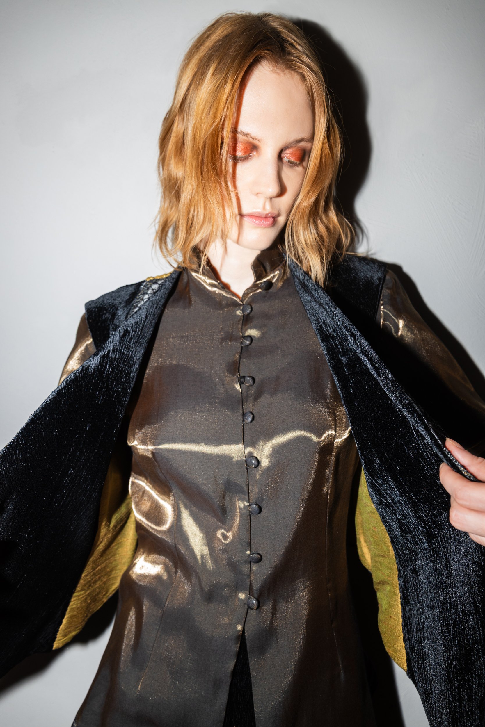 A woman with shoulder-length reddish hair, wearing metallic gold and black clothing, with her eyes closed, holding open a black and gold jacket.
