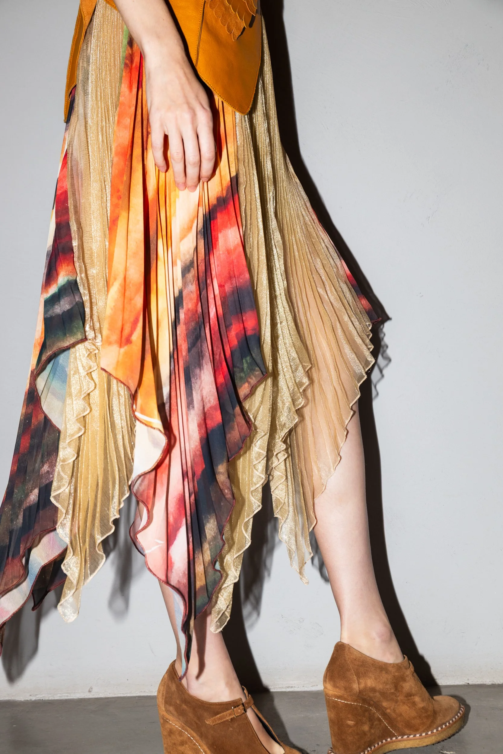 Part of a woman wearing a flowing, pleated midi skirt with an abstract, colorful pattern, paired with brown suede wedge heels, standing against a plain wall.
