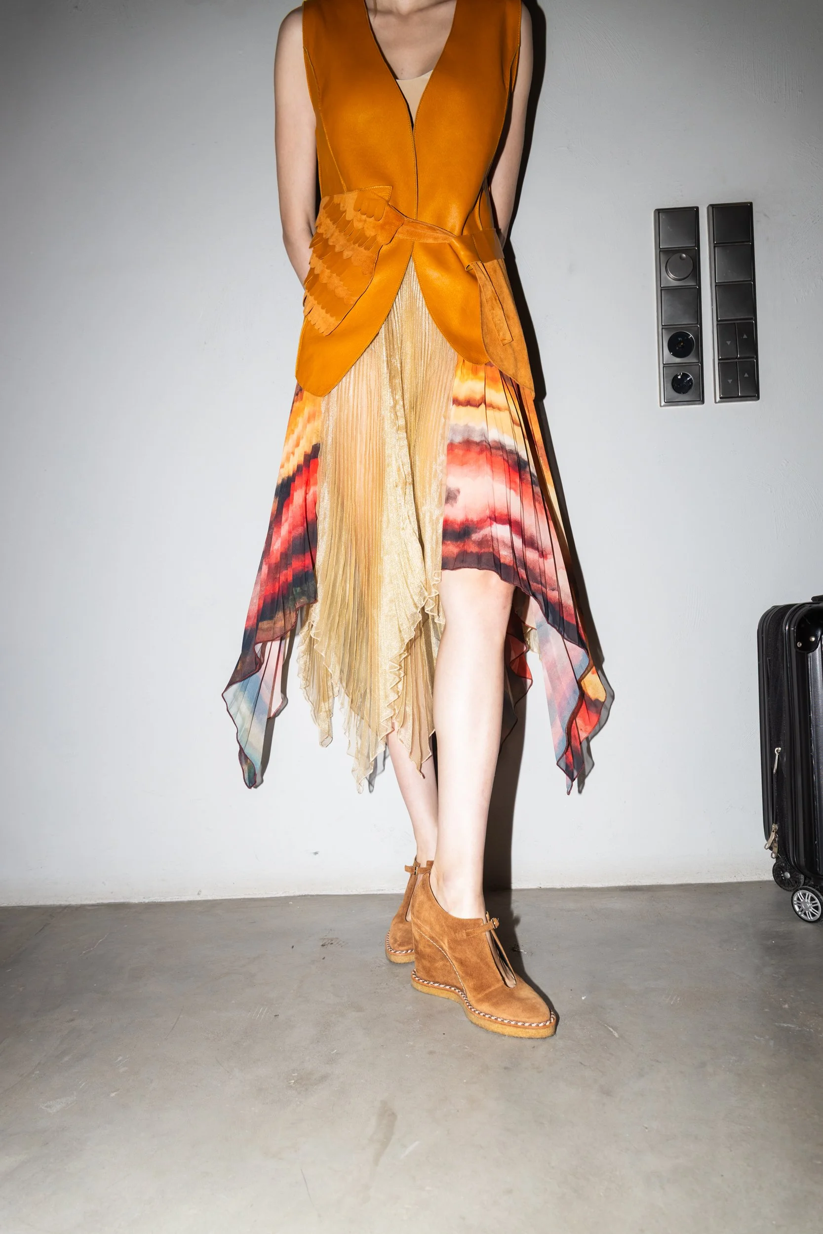 Model standing against a wall in a sleeveless orange top, layered multicolored skirt, and tan ankle boots.