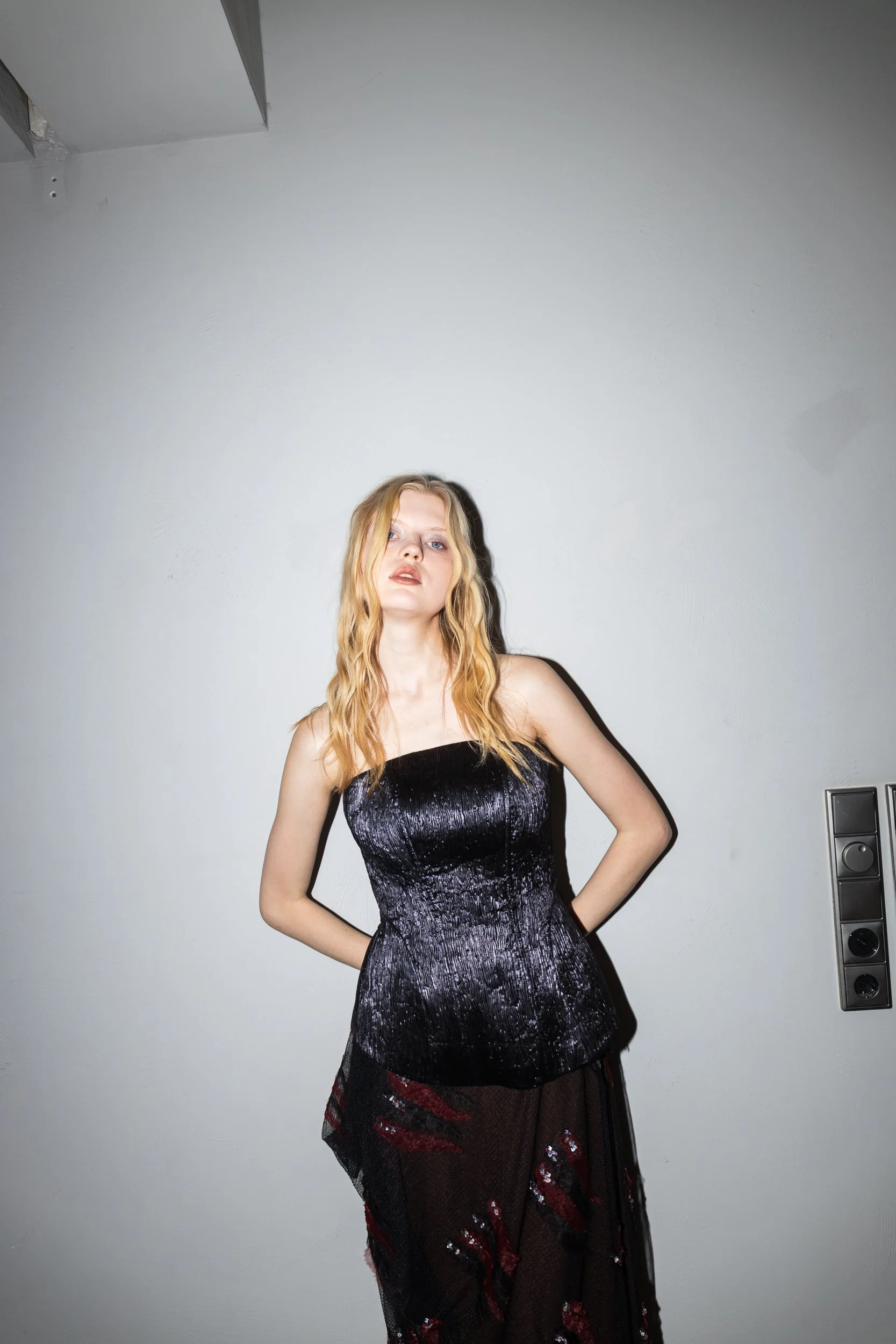 A young woman with blonde hair and fair skin posing in front of a plain wall. She is wearing a strapless black dress with a shiny texture and a sheer black skirt with red accents. Her left hand is on her hip, and her right hand is behind her back. Th