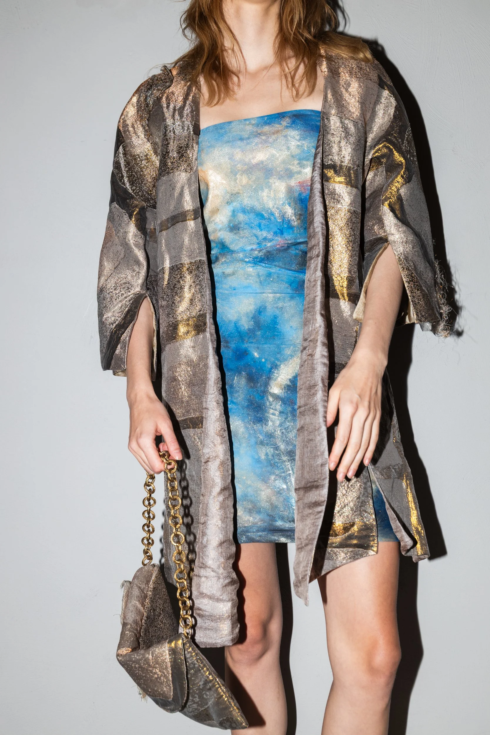 A woman wearing a strapless blue and gold abstract dress, layered with a metallic gold and black patterned robe, holding a matching metallic gold and black purse with a chain strap.