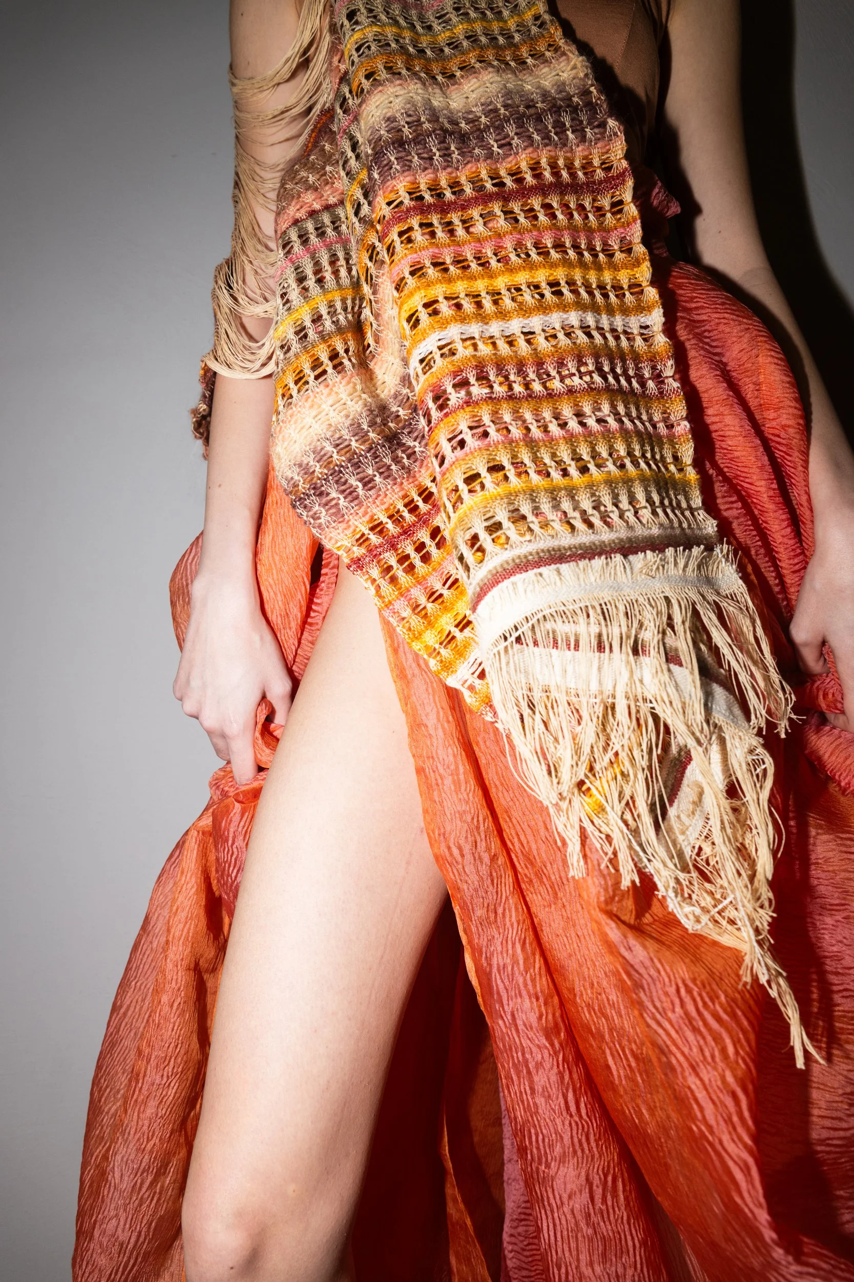 A person wearing a colorful, textured scarf with fringe details and a flowing, sheer orange skirt that reveals their thigh.