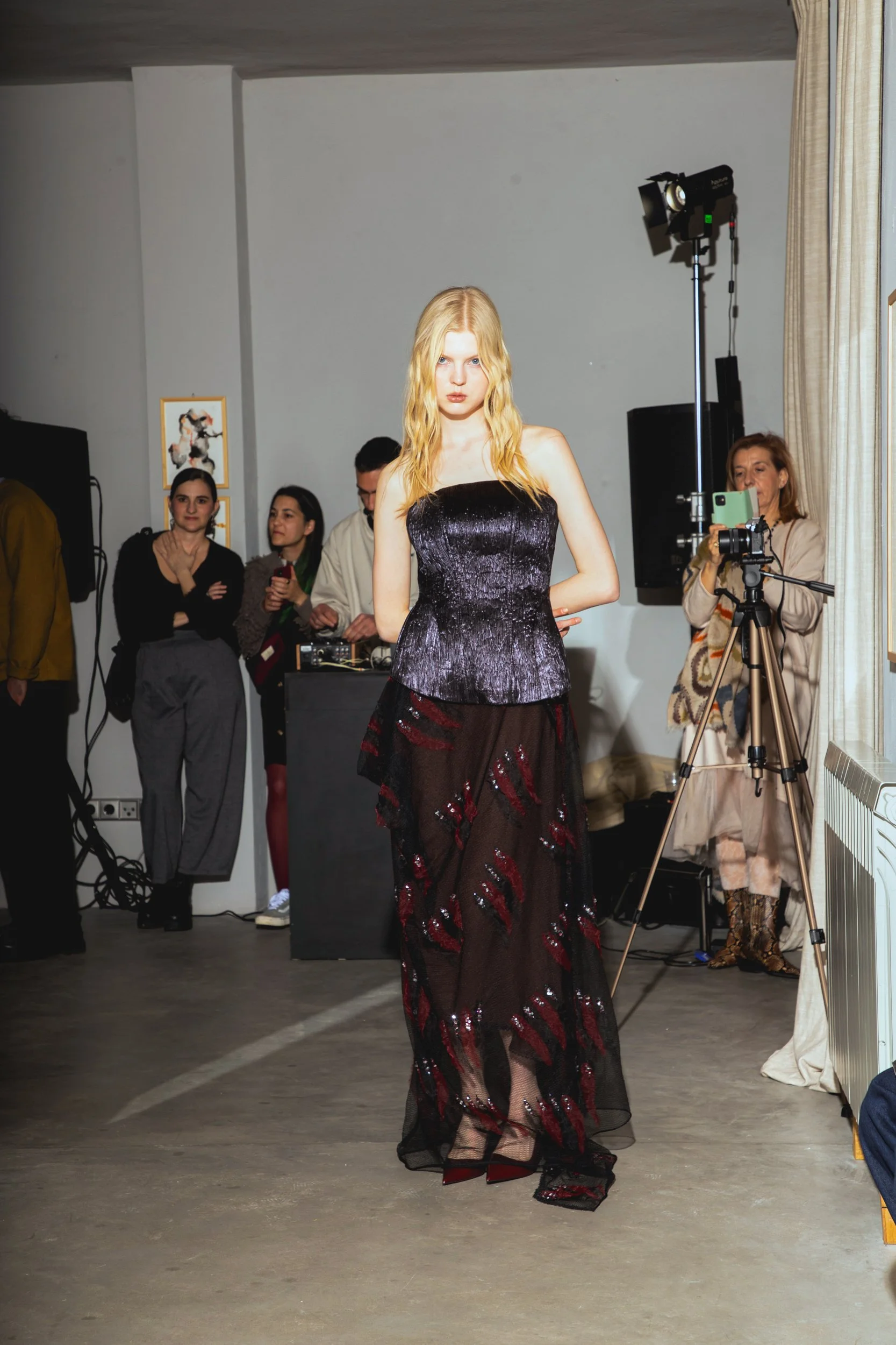 A fashion model in a black and red sequined dress walking down the runway during a fashion show, with onlookers and photographers in the background.