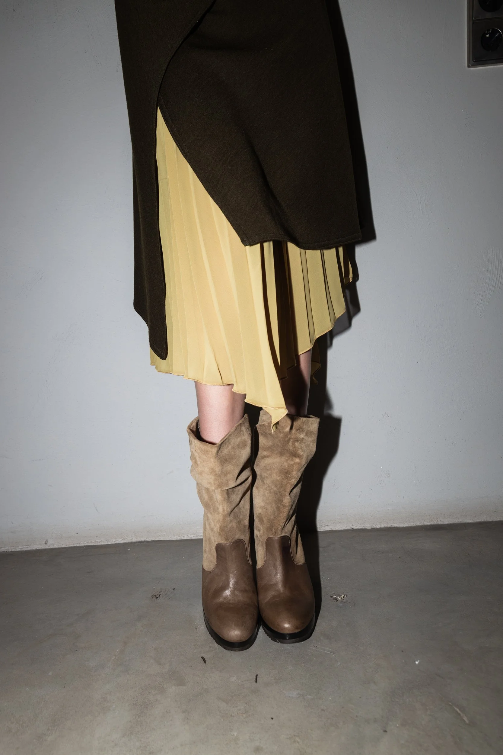 Person wearing beige leather and suede boots, a black coat, and a yellow pleated skirt, standing against a plain white wall and cement floor.