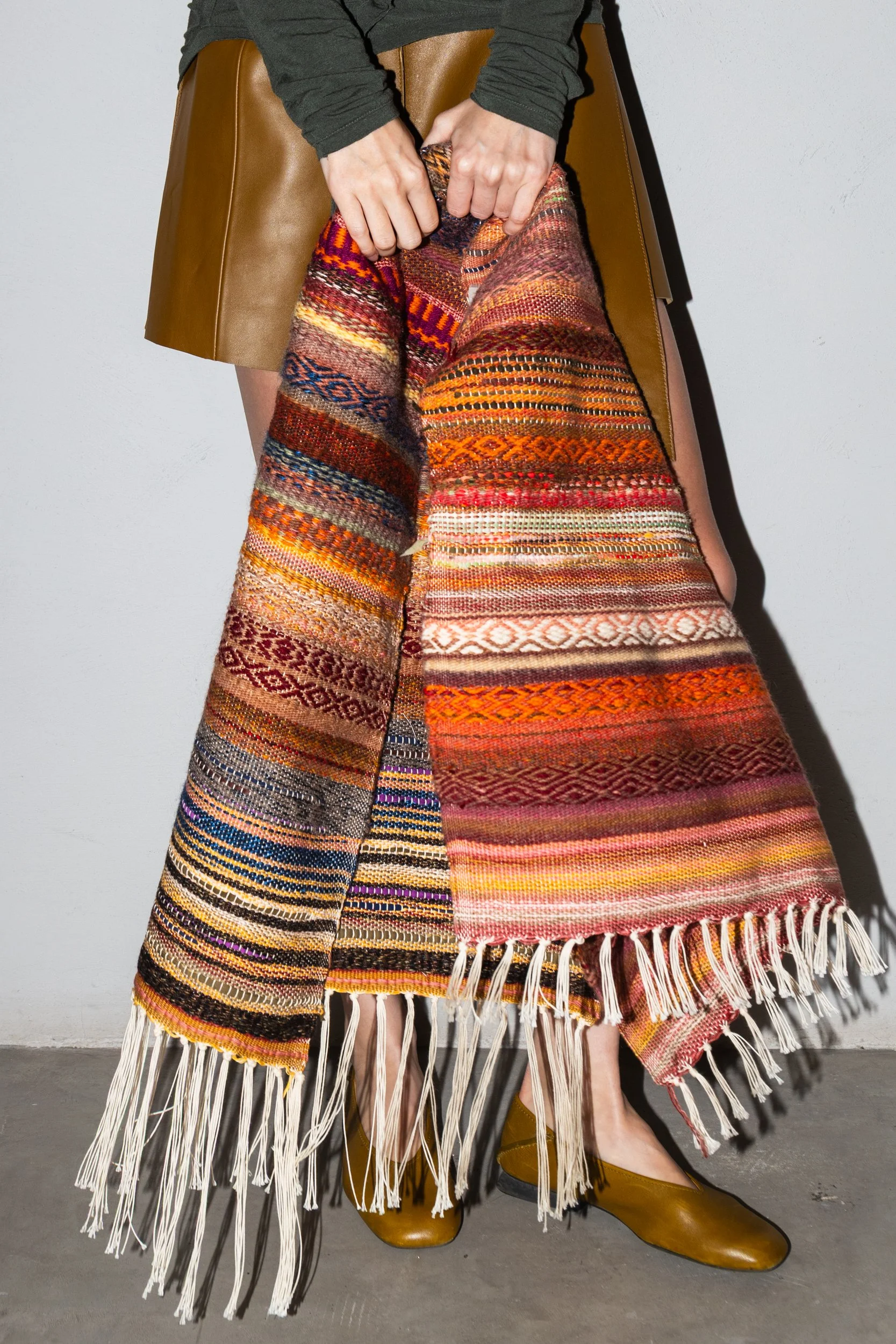 Person holding a colorful woven blanket with fringes, wearing brown shoes and a brown skirt, standing against a light gray wall.