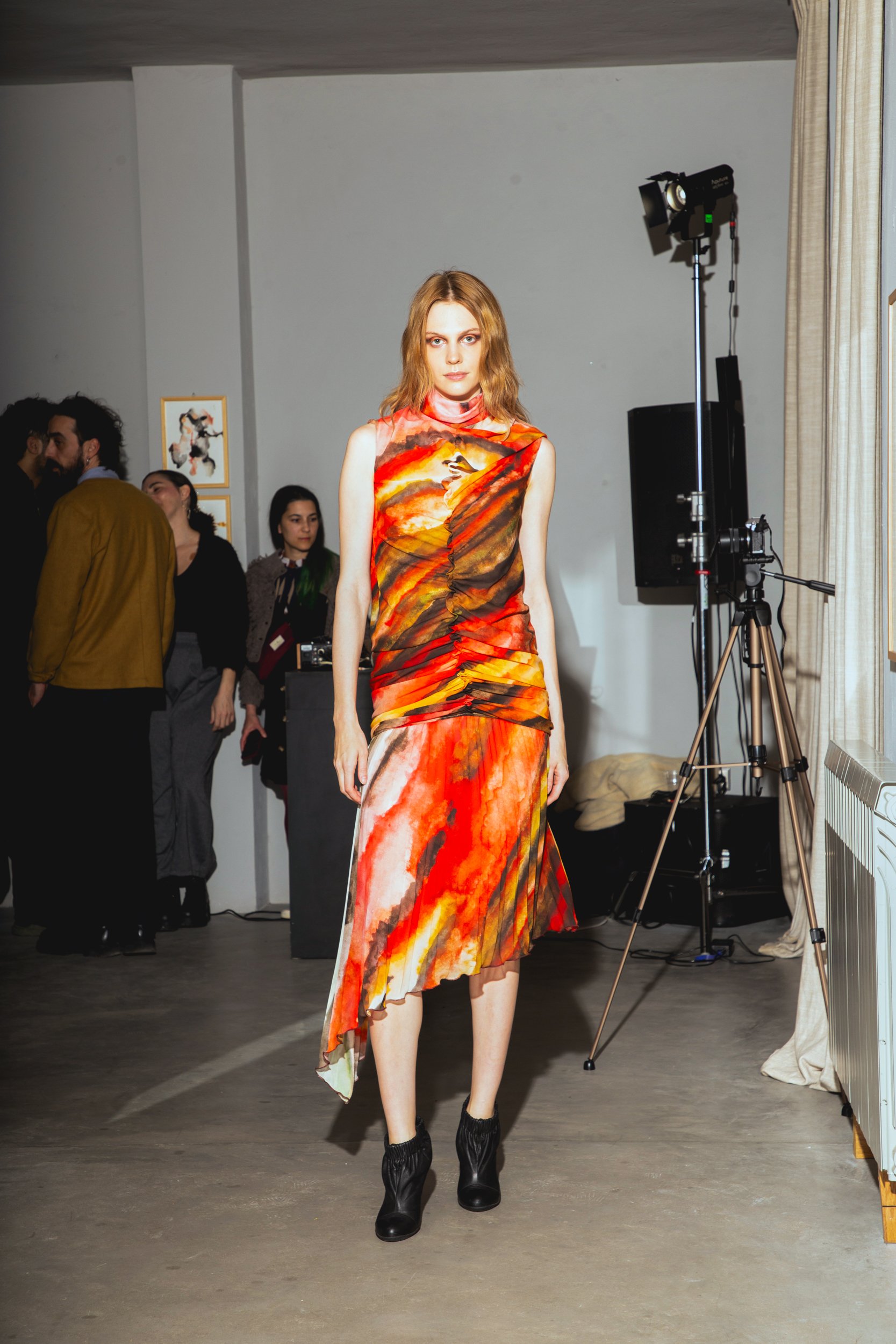 A female model walking on a runway in a vibrant red, orange, and black watercolor-style dress paired with black ankle boots, at a fashion show or art event.