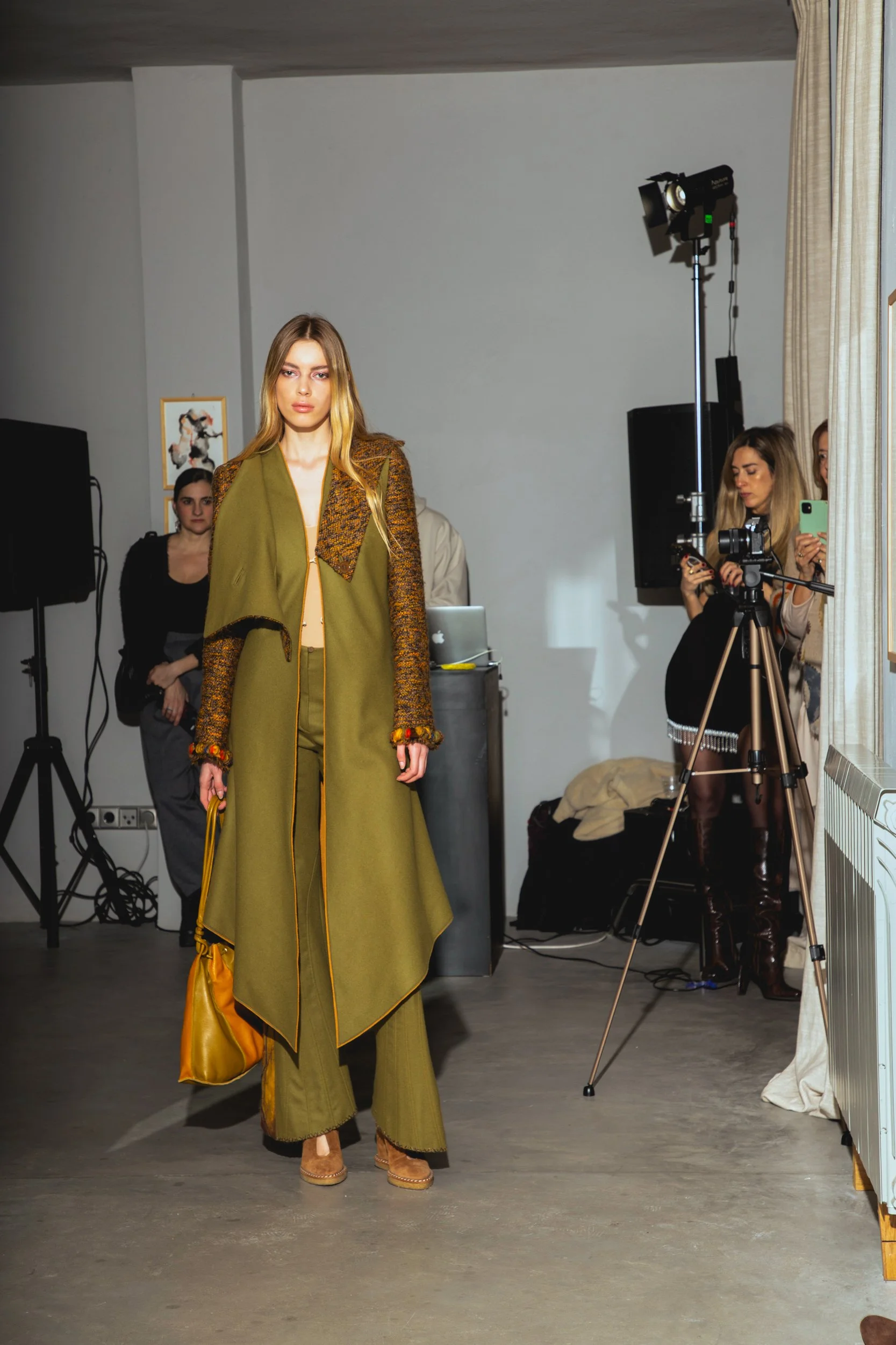 Model walking on runway during fashion show with photographers and audience in background.