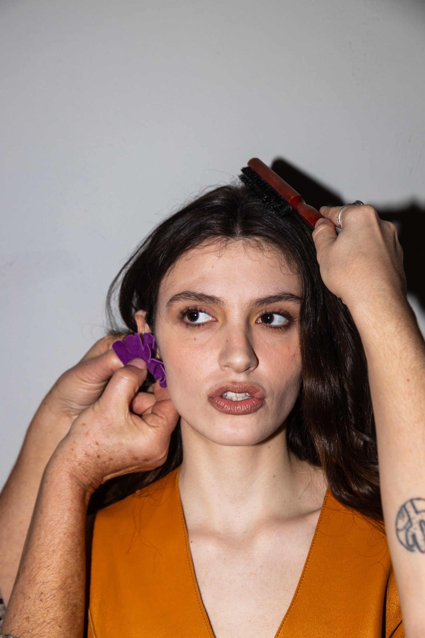 A woman with long dark hair getting her hair styled, with a person using a hairbrush on her hair and holding a purple hair accessory, against a plain white background.
