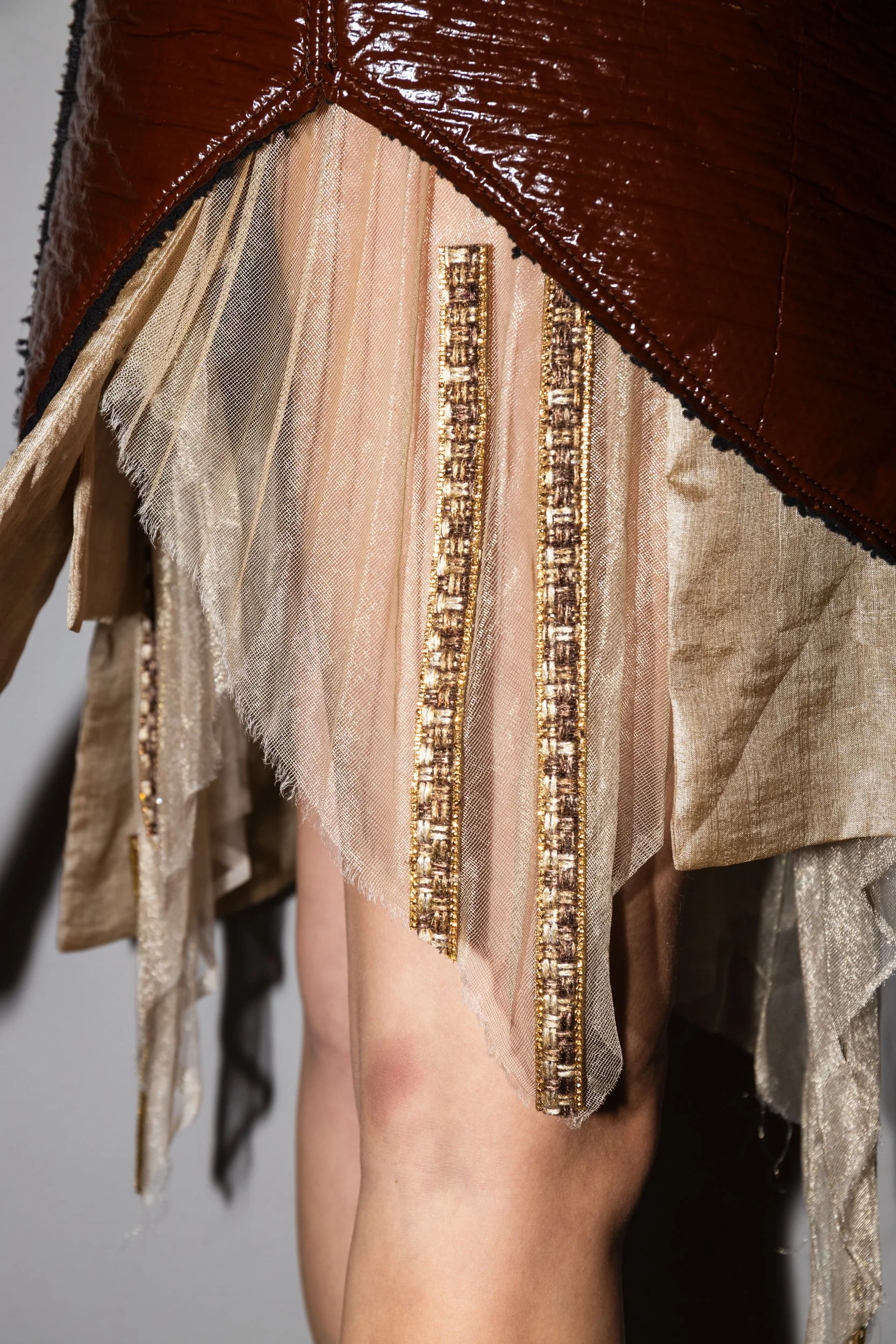 Close-up of a model's leg wearing a metallic gold fabric dress with sheer beige fabric and gold-embellished strips, partially covered by a brown leather jacket.