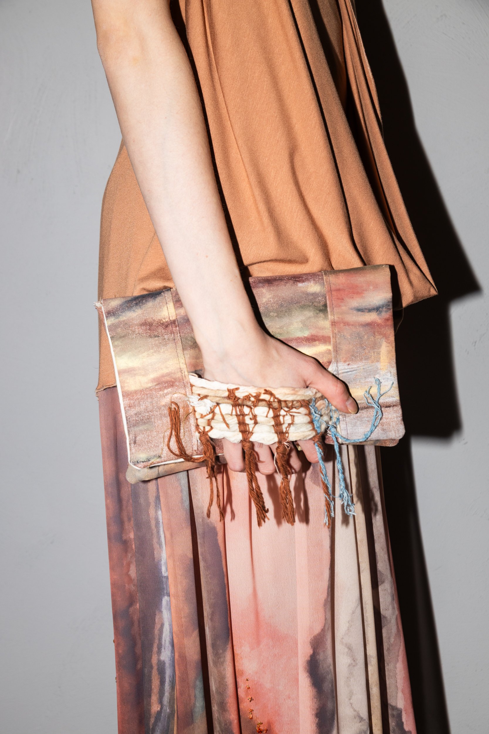 A person is holding a clutch bag with a landscape painting on it, wearing a tan outfit with black piping accents.