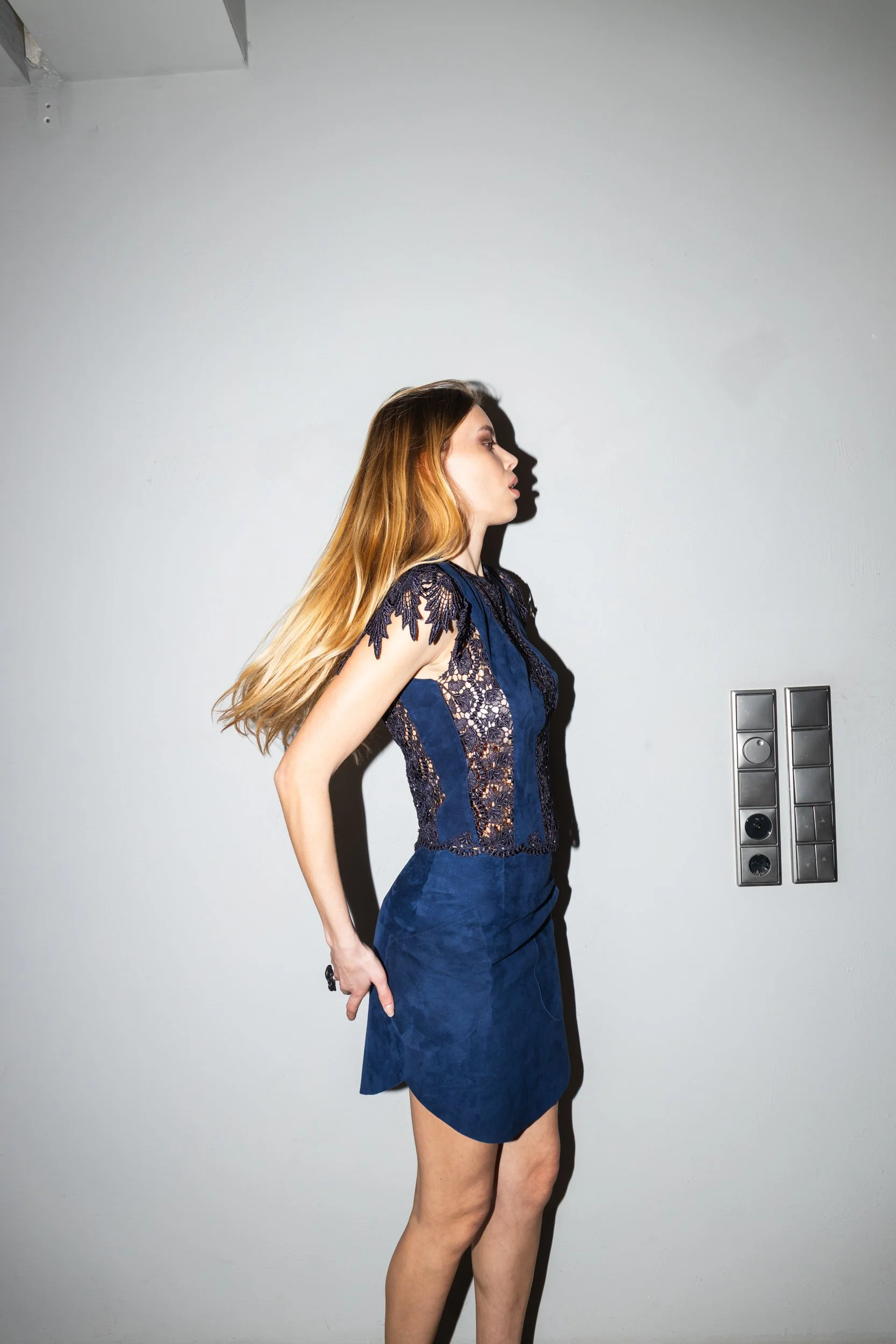 A woman with long, blonde hair wearing a blue dress with lace detailing, standing against a plain light-colored wall.