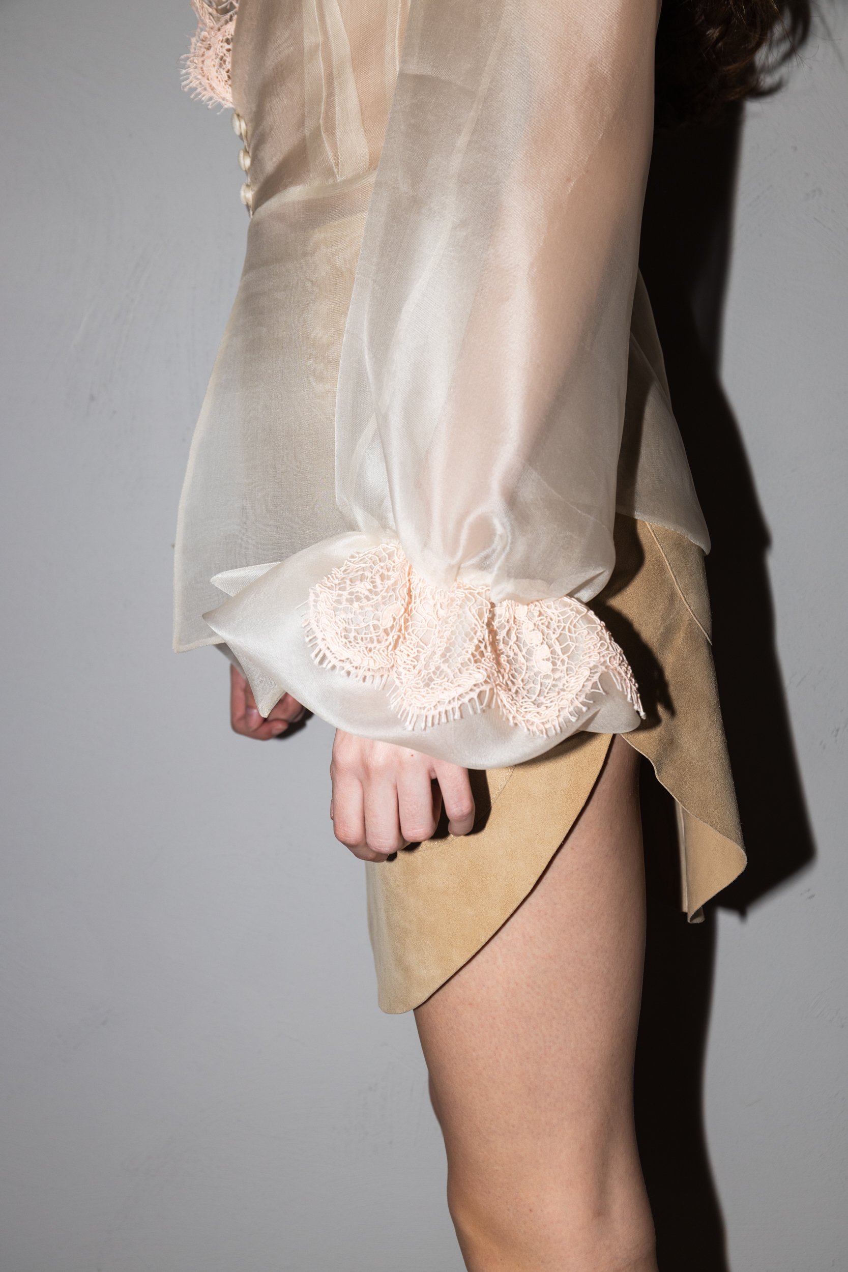 Close-up of a person wearing a sheer, beige, long-sleeve top with lace detail on the sleeve cuffs and a beige miniskirt, standing against a plain wall.