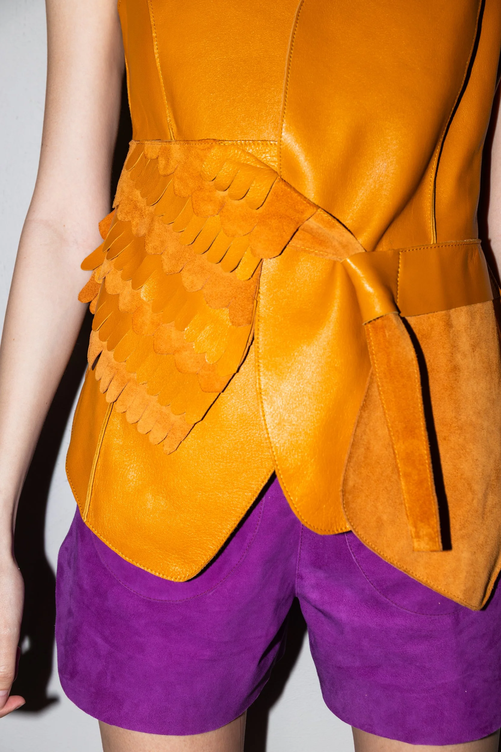 Close-up of a person wearing a bright orange sleeveless leather top with leaf-shaped embellishments and purple shorts.