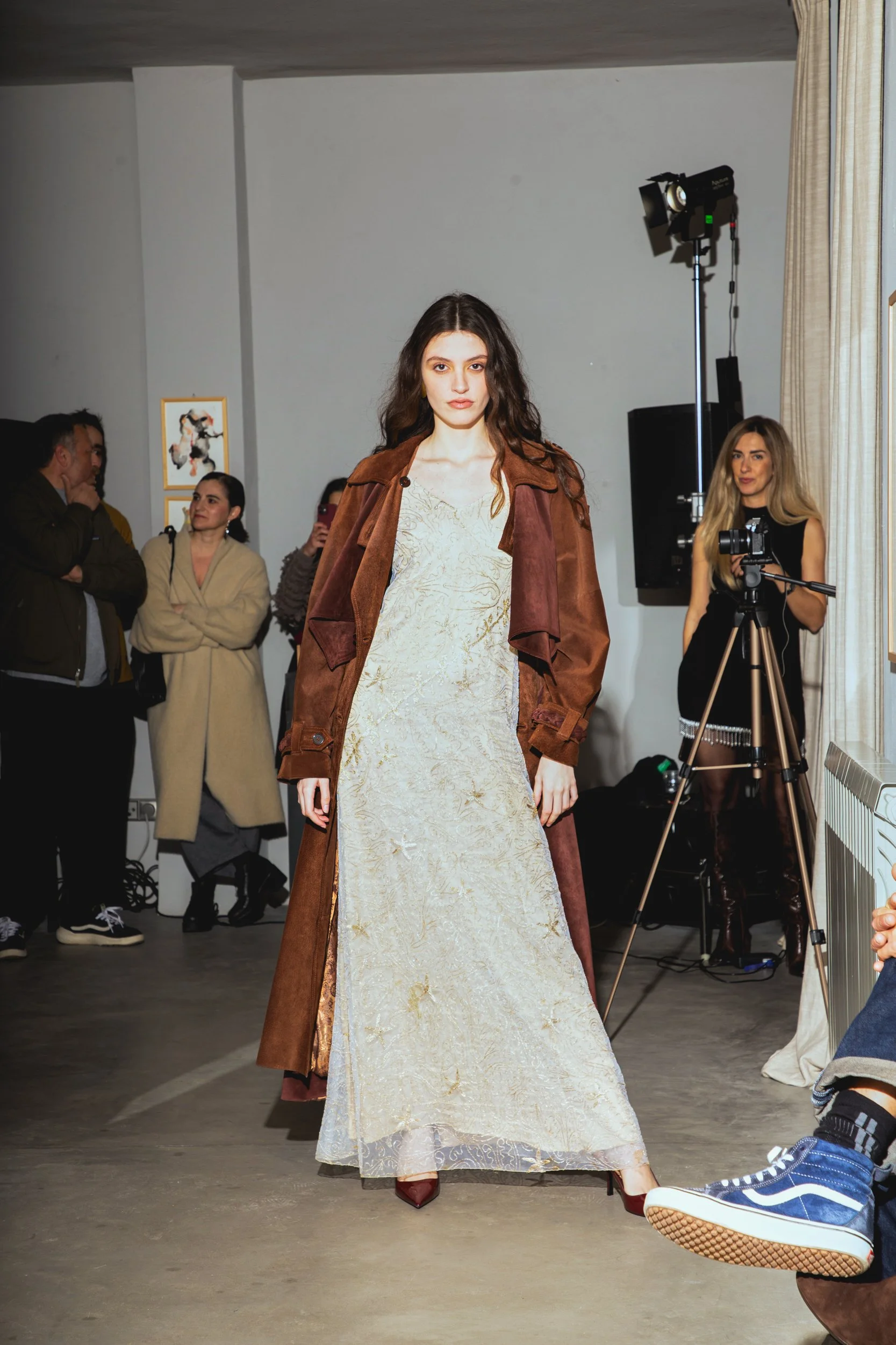 A female model walking on a runway during a fashion show, wearing a cream-colored dress with gold embroidery and a brown trench coat, with spectators and a photographer in the background.