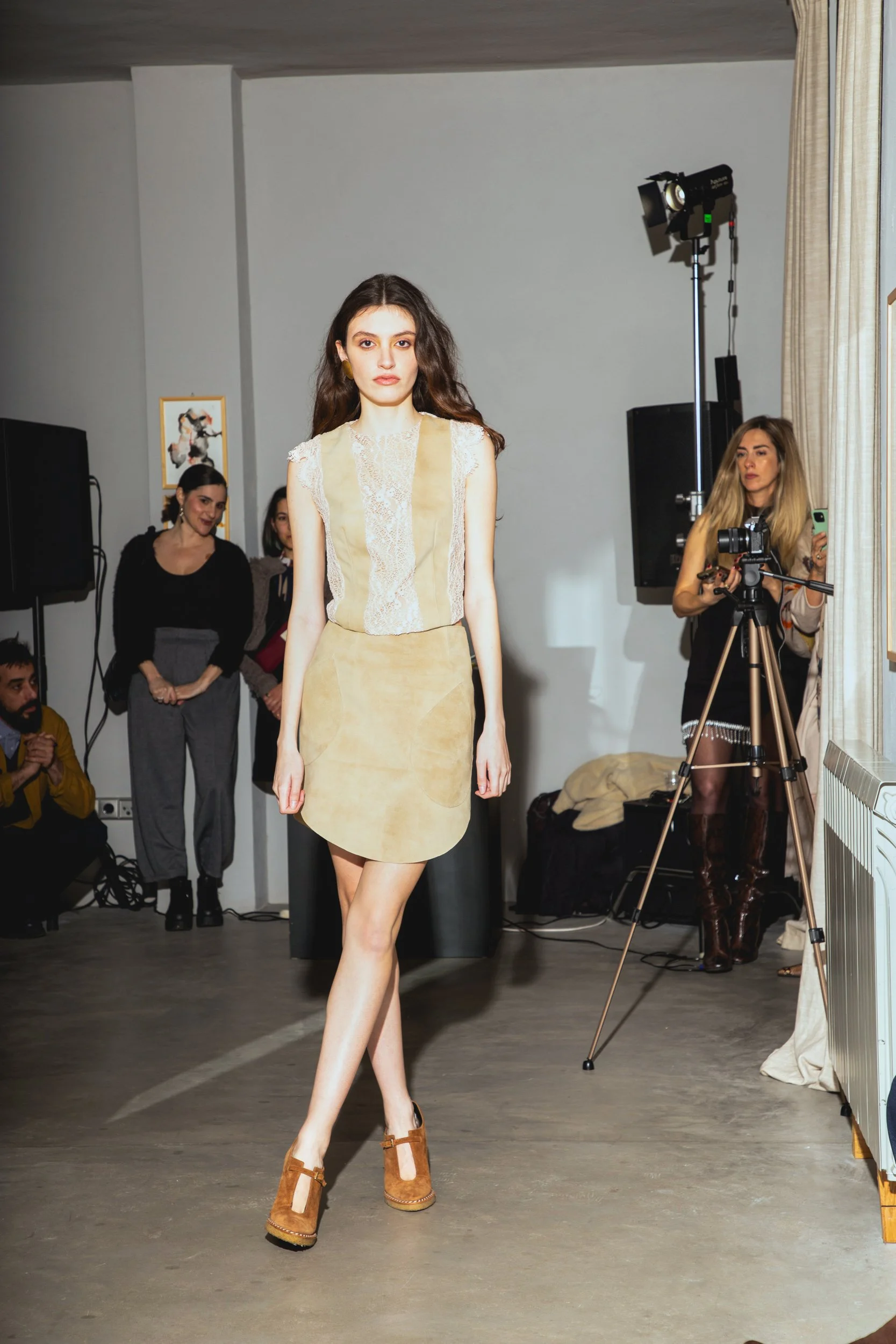 A young woman walks on a runway during a fashion show, with a camera and crew filming her, and spectators watching.