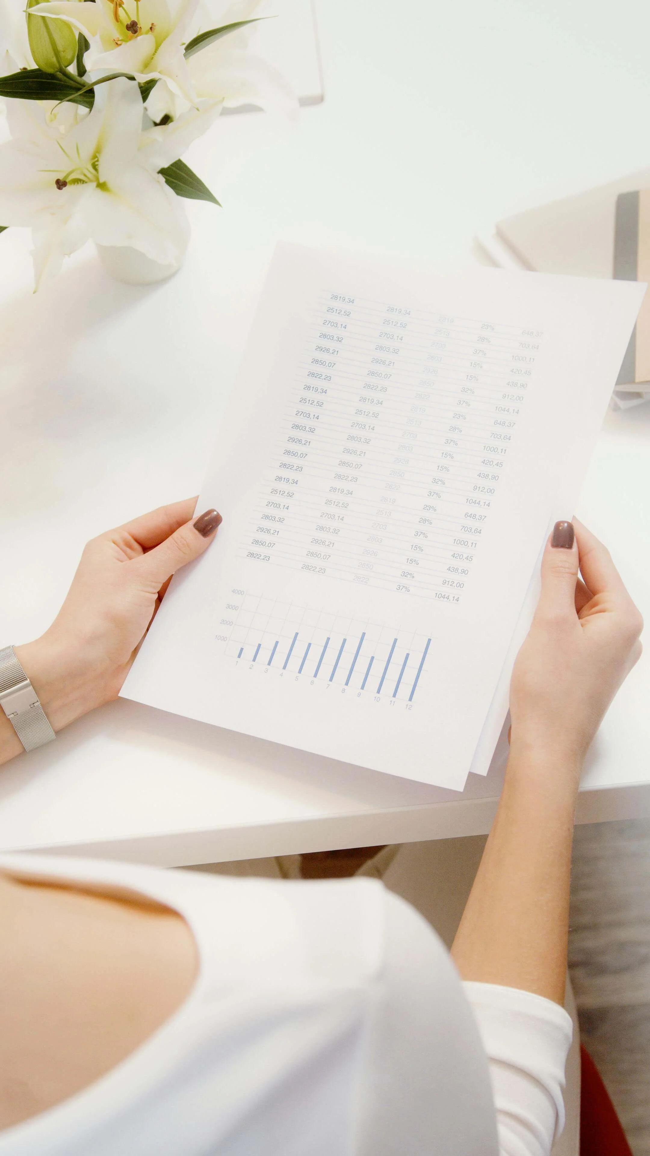 Close-up of female hands in white blouse gripping a printed financial document showing data charts, next to flowers on white table.