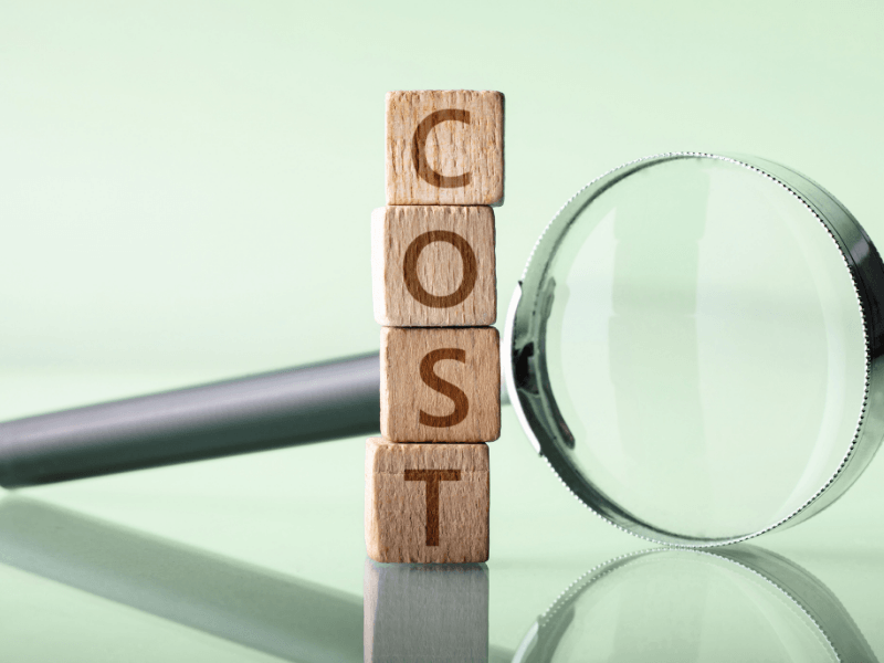 A magnifying glass examines wooden blocks that spell out the word cost, a key factor when planning retirement savings.