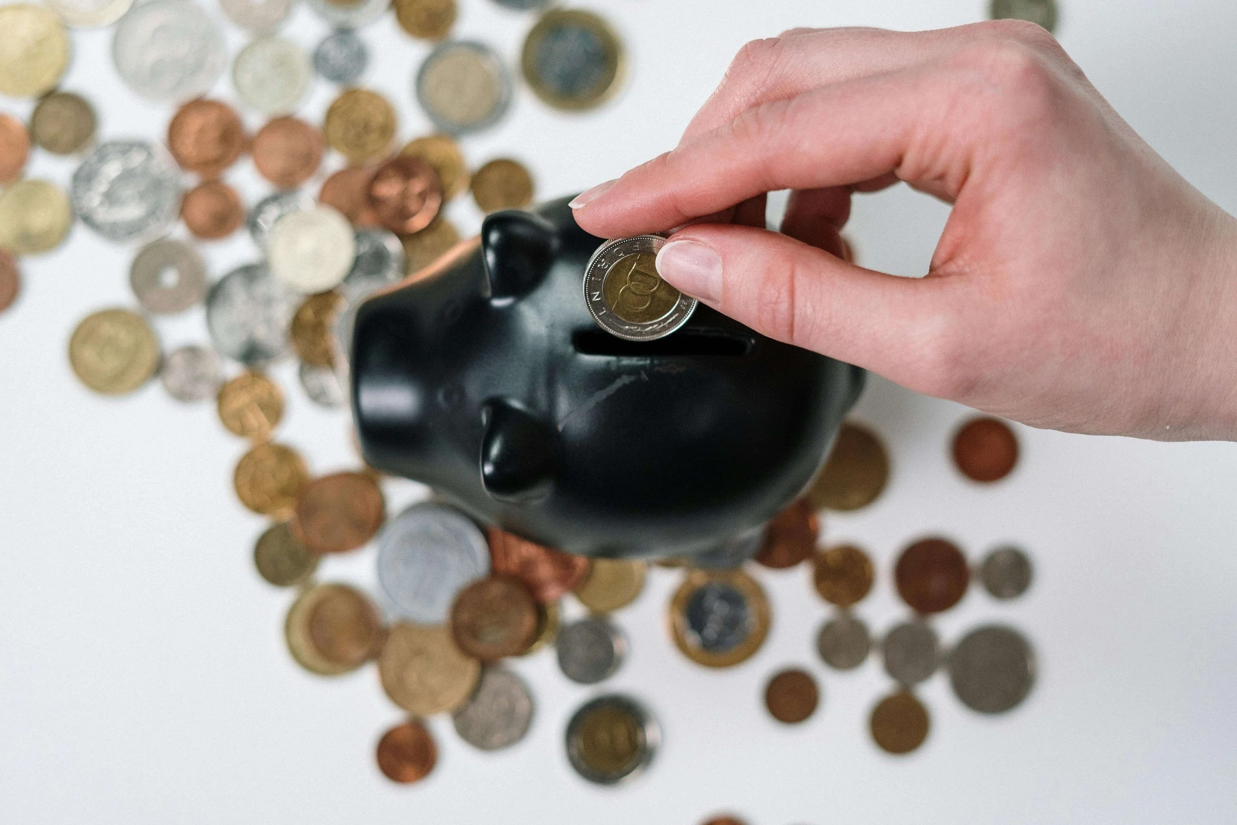 Close-up of a black piggy bank with coins spilling out and a hand inserting another coin to build savings.