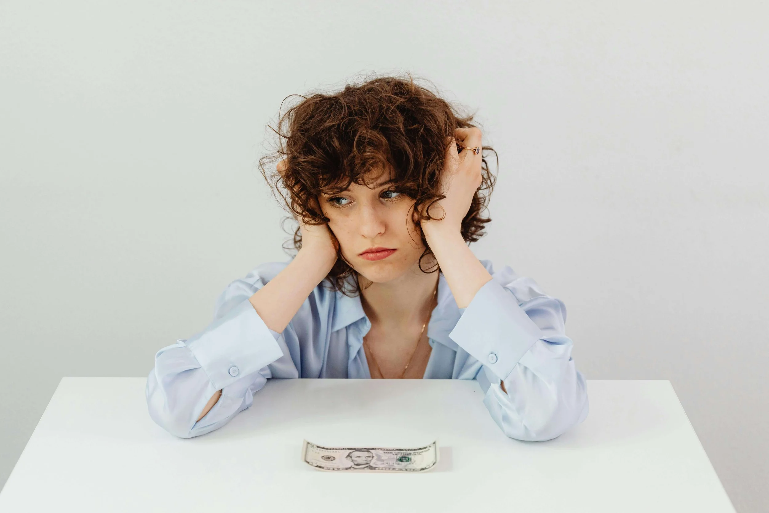 Frustrated woman in light blue shirt with hands on head, staring at a single dollar bill on table during money-saving challenge.