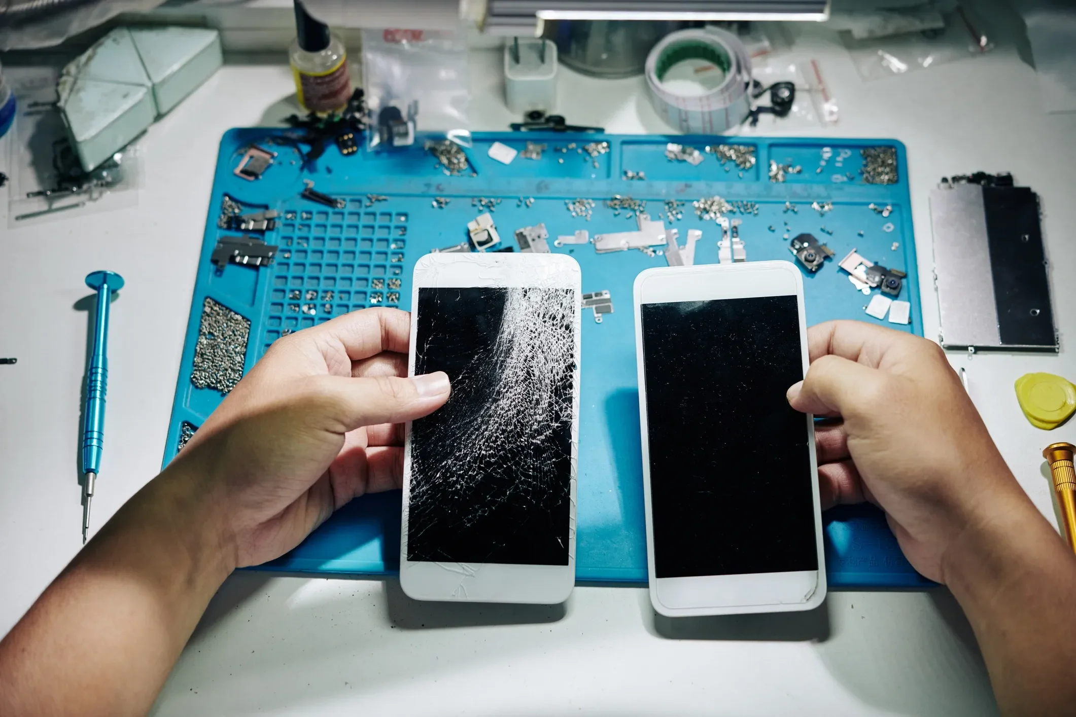 Two damaged smartphones with cracked screens on a workspace with various disassembled electronic parts and tools.