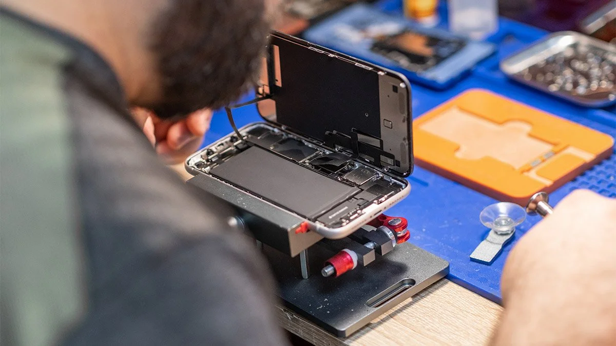 Person repairing a smartphone with its screen removed, working at a workbench with repair tools and spare parts.
