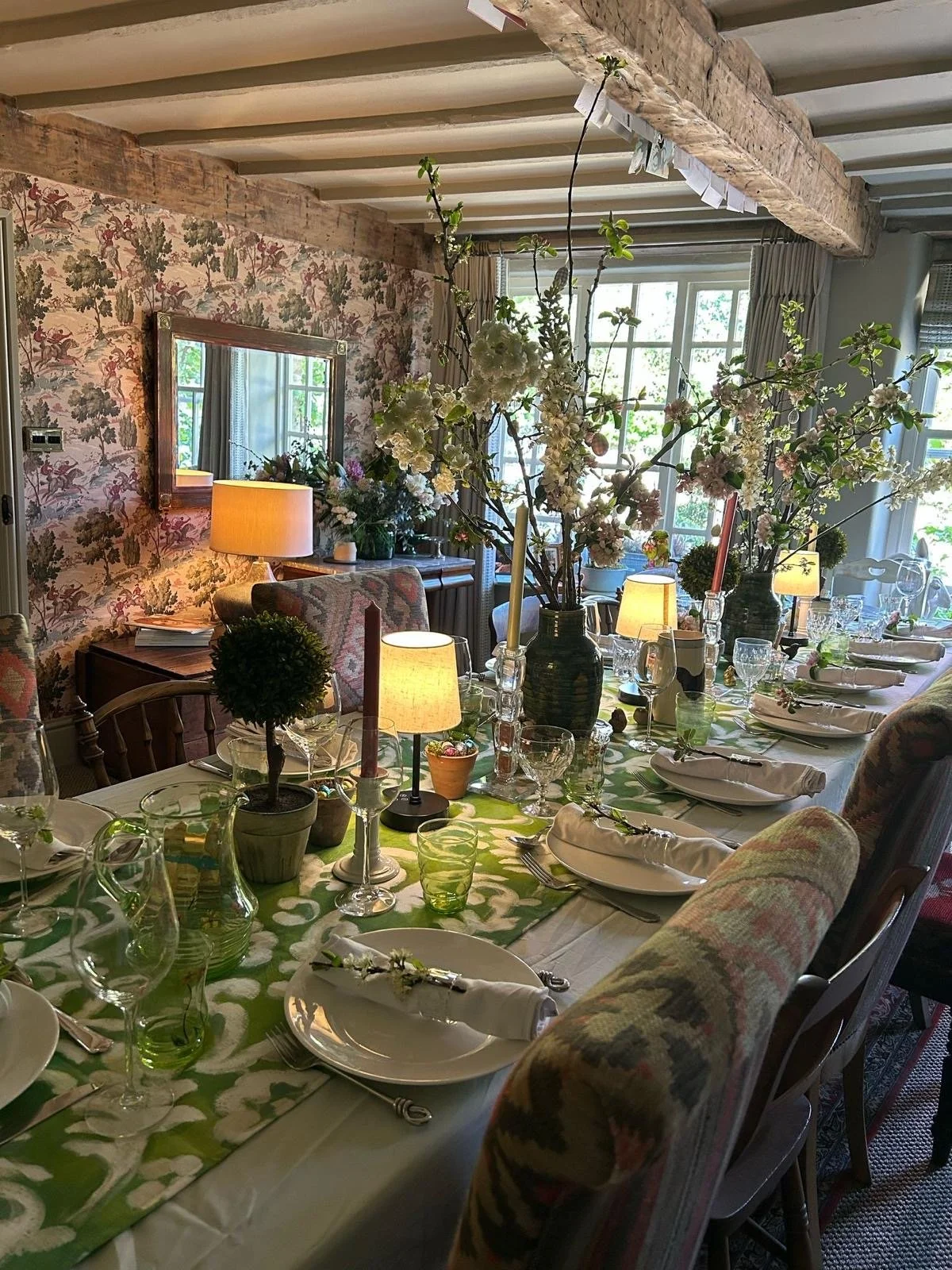 A dining table laid with white tablecloth, green table runner, candles, lamps, foliage, and linen napkins.