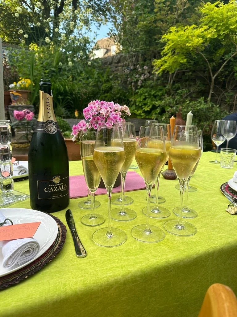 A bottle of Champagne and filled flutes sit on a dining table laid with a green tablecloth, candles, pink flowers, and linen napkins.