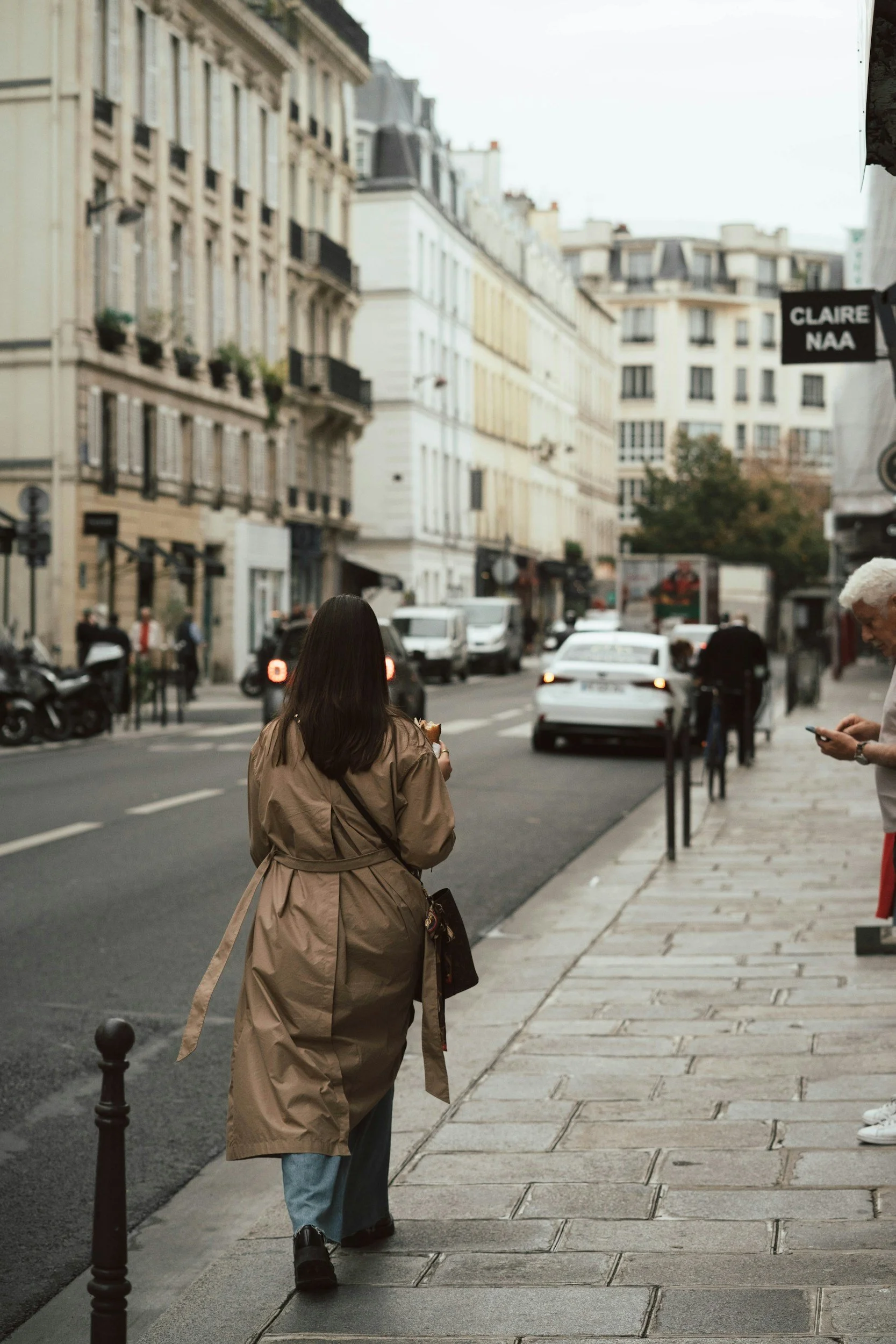 A young woman with dark hair wearing a beige trench coat and blue jeans walking on a city sidewalk, with cars driving down the street and older style apartment buildings in the background.