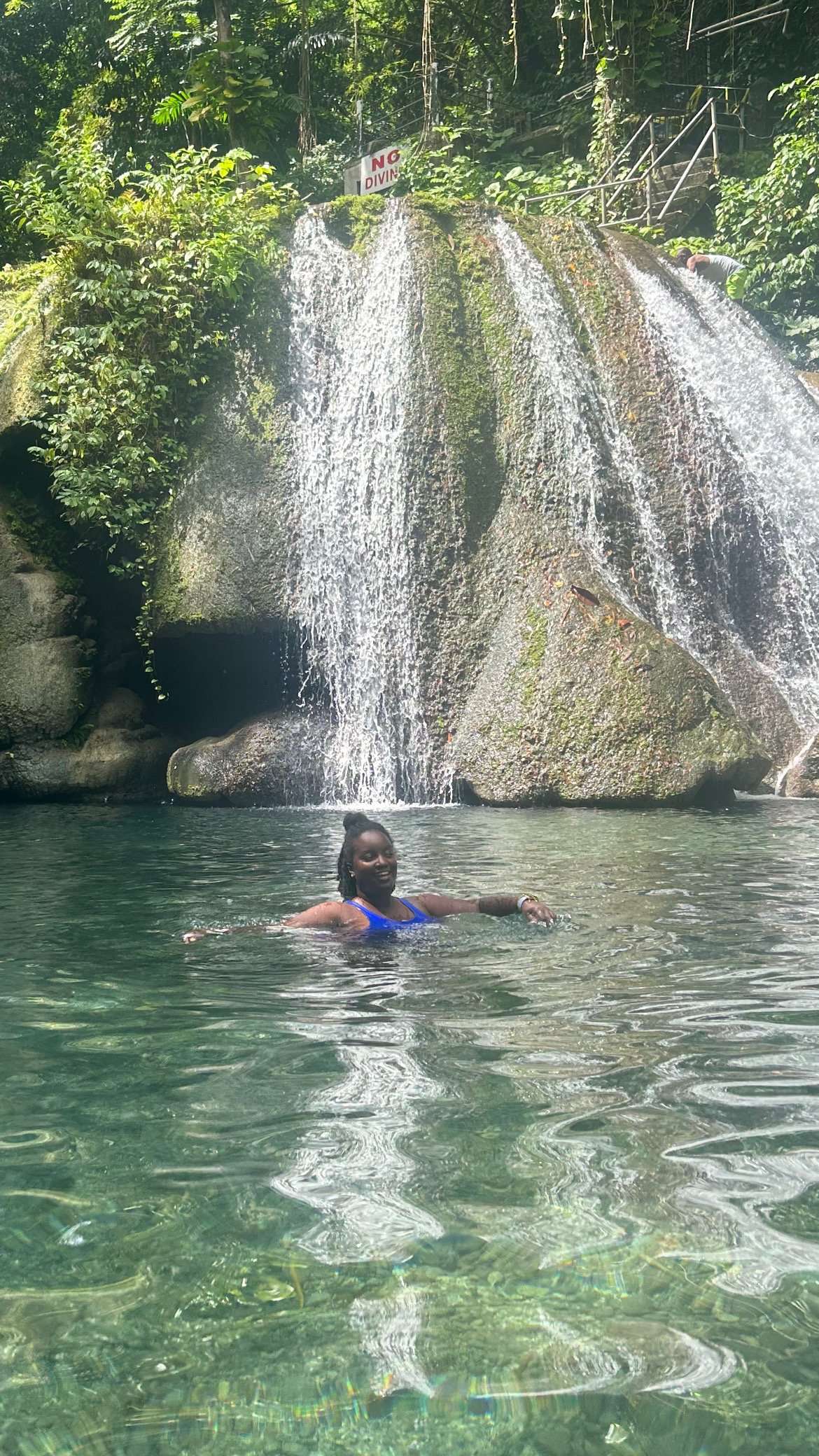 A woman swimming in a natural pool at the base of a waterfall surrounded by lush green trees.